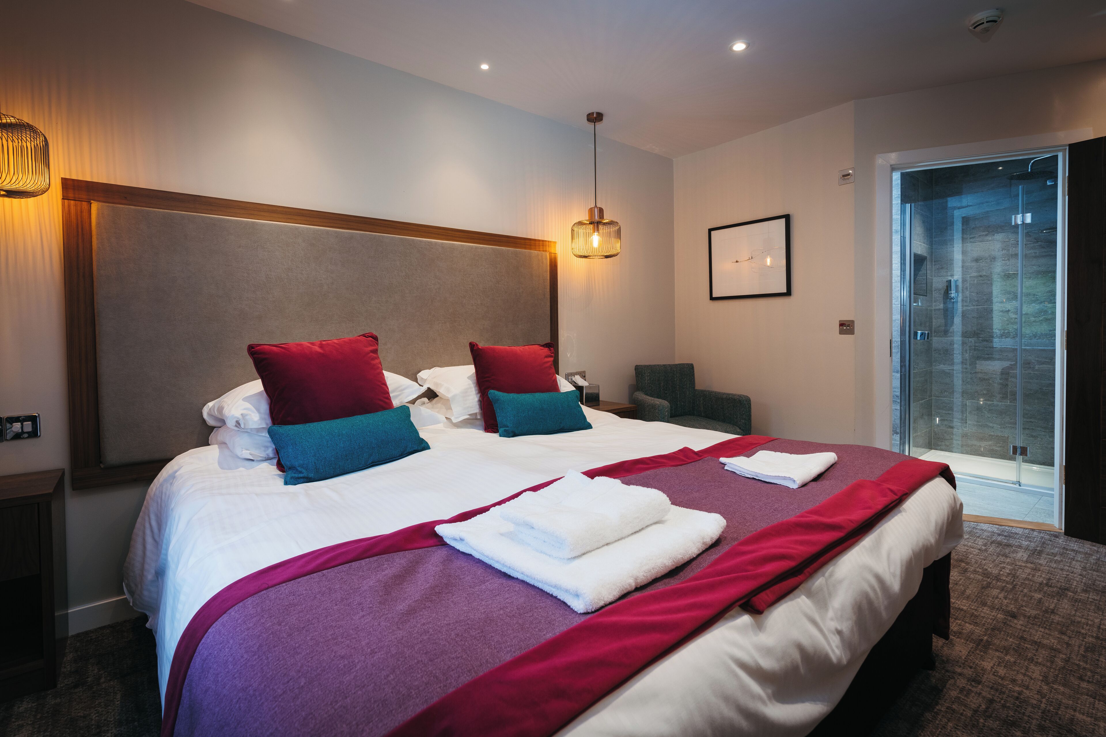 Habitación de hotel moderna con una cama grande, dos almohadas rojas y dos azules, una colcha roja y blanca, y una puerta de ducha de cristal.