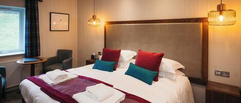 A hotel room with a large bed, two bedside tables, a chair, a small table, and a window with curtains.