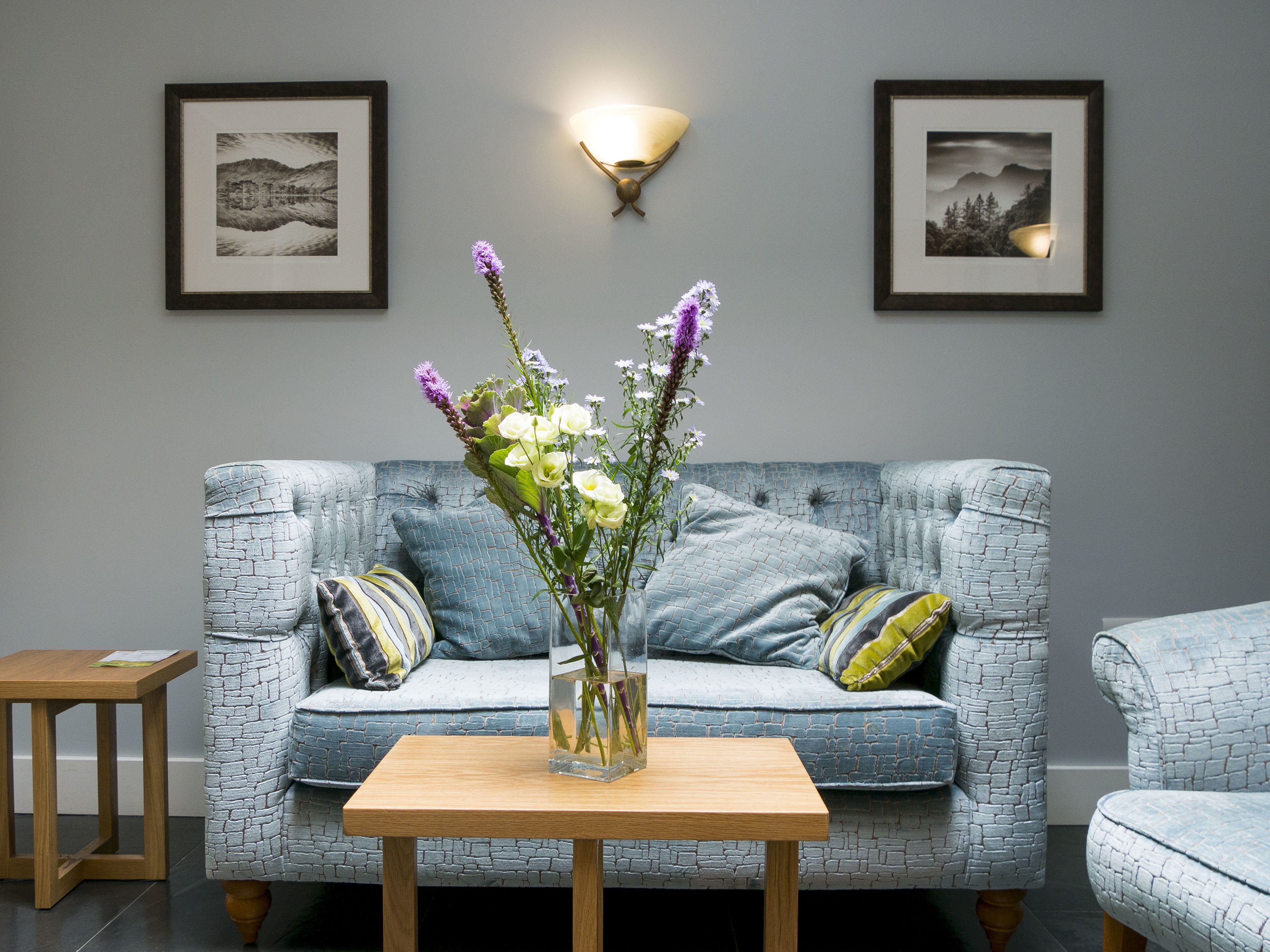 Una sala de estar con un sofá azul con textura, una mesa de centro de madera y un jarrón con flores. Hay dos cuadros en la pared y un aplique de pared.