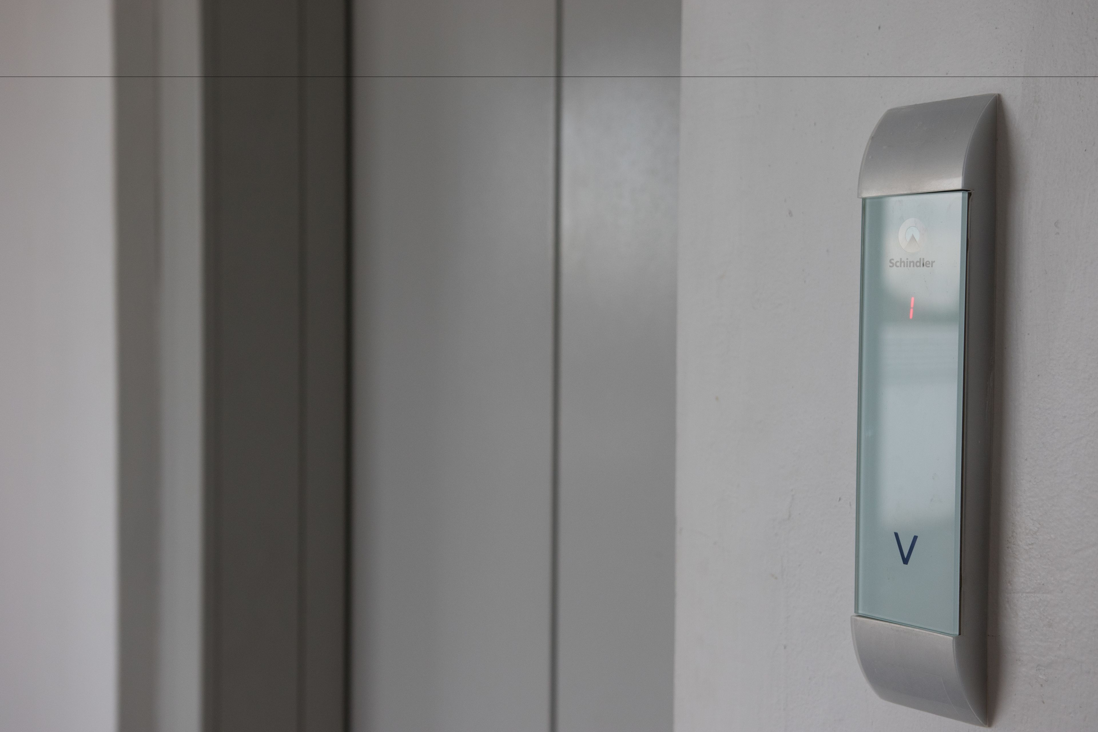 A modern elevator with a minimalist design, featuring a glass panel and a control panel with a 'V' button.