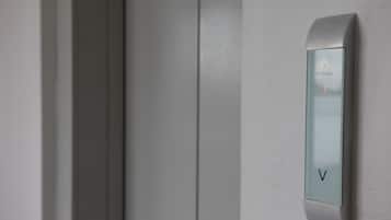 A modern elevator with a minimalist design, featuring a glass panel and a control panel with a 'V' button.