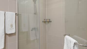 A glass-enclosed shower with a handheld showerhead, a wall-mounted faucet, and a shelf with toiletries.
