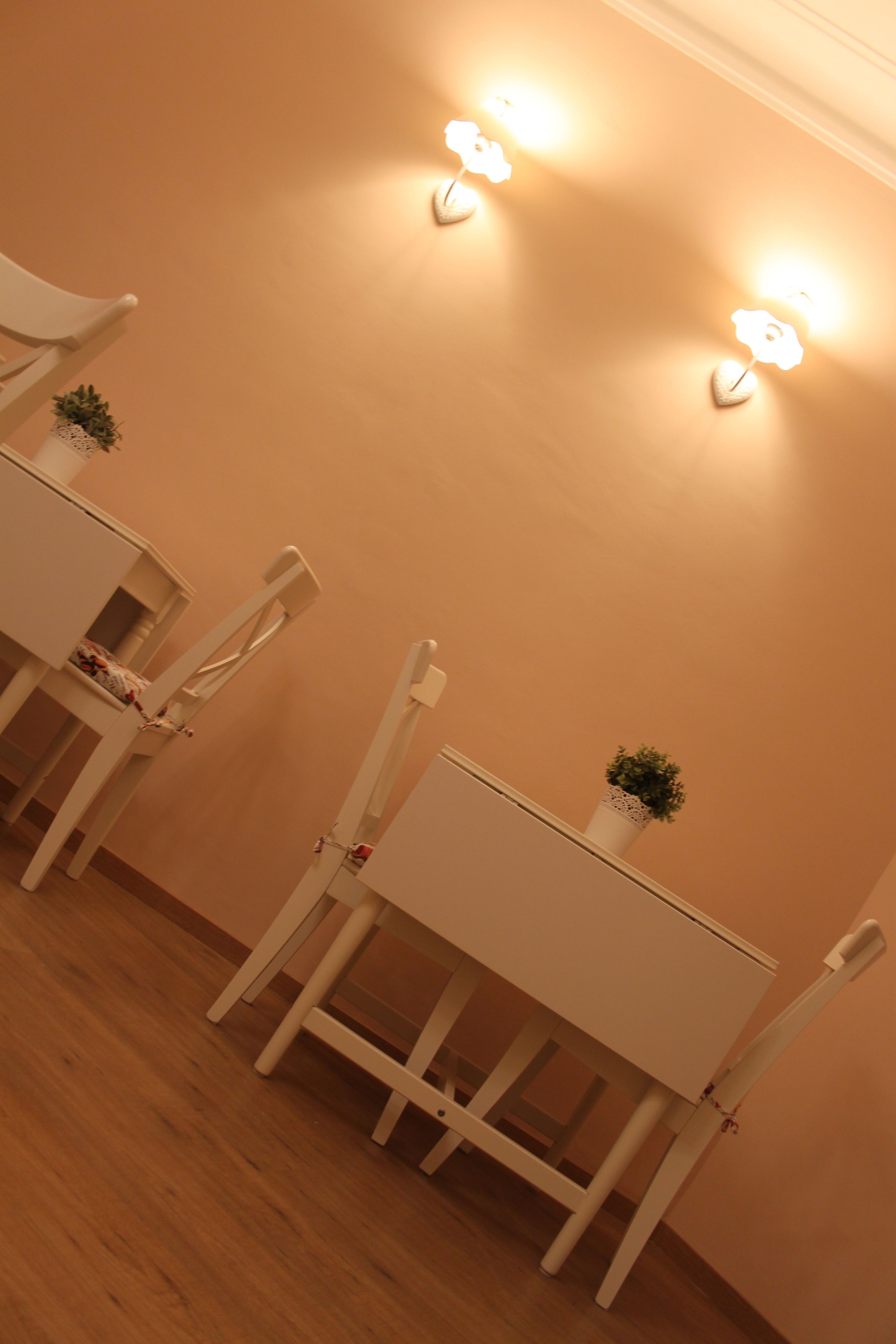 A dining area with white tables and chairs, two wall-mounted light fixtures, and a small potted plant on one table.