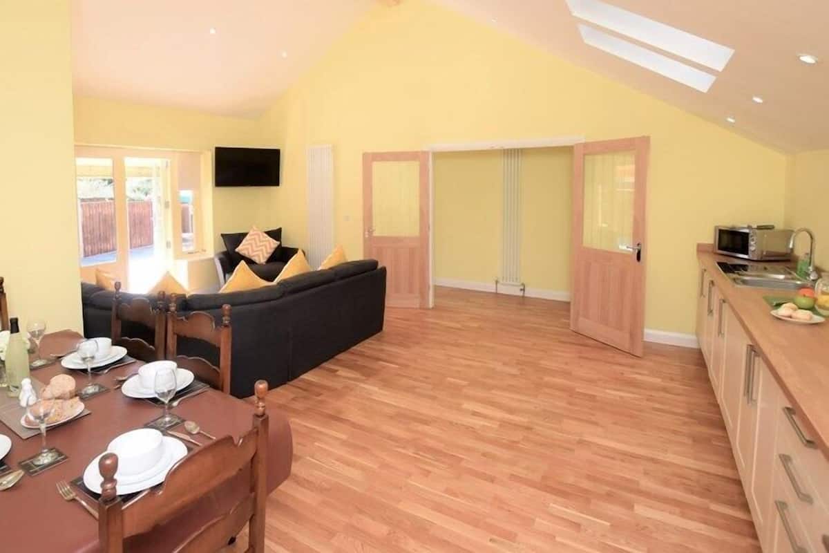 A spacious living area with a wooden dining table set for a meal, a black sofa with cushions, and a kitchen area with a microwave and sink.