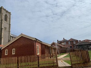 A brick house with a wooden fence, a grassy yard, and a church in the background. - The White Lady (North Walsham)