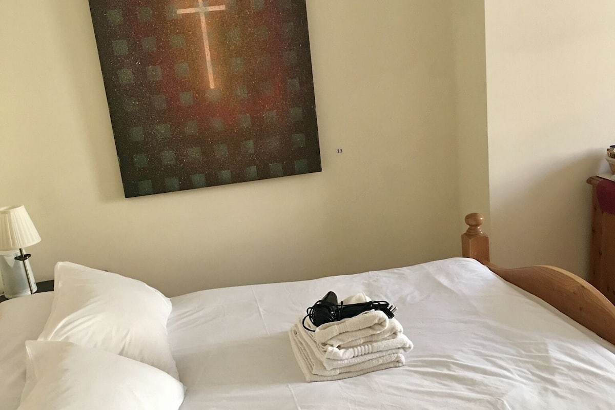 A neatly made bed with white linens, a folded towel, and a pair of glasses on the bed. A cross-shaped artwork is hung on the wall above the bed.