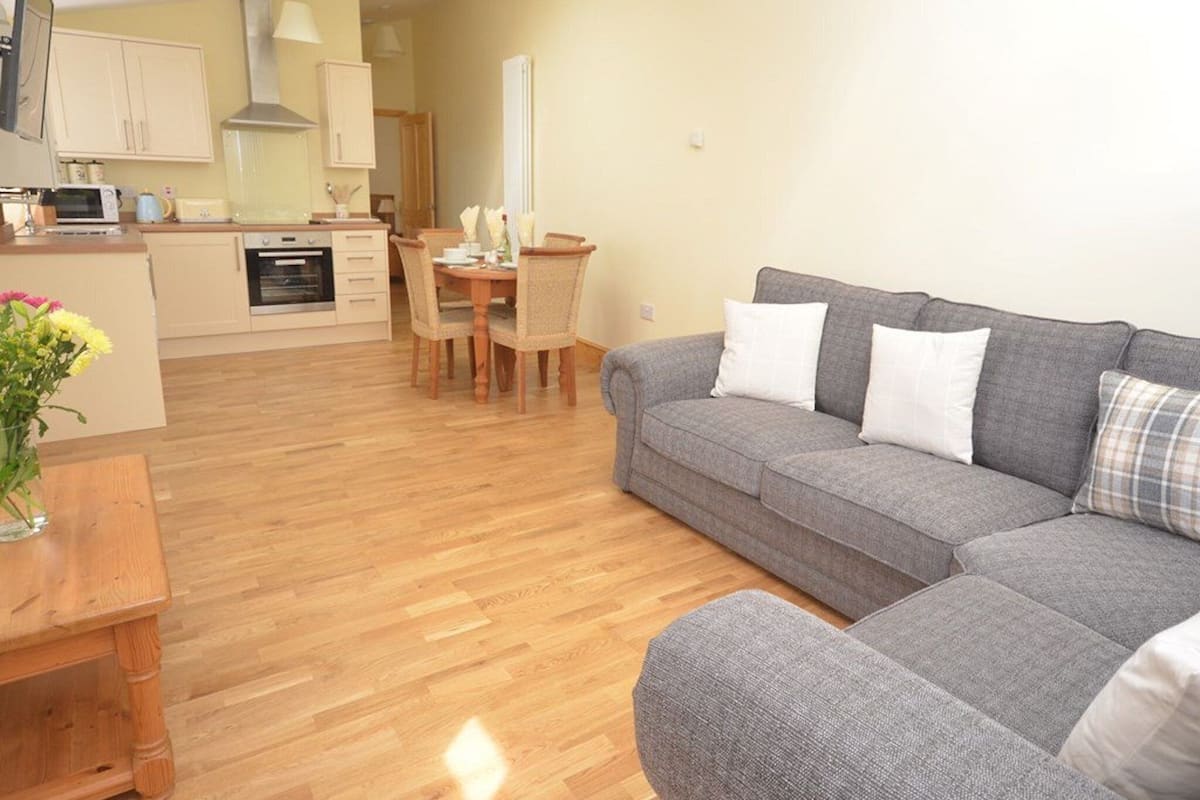 A modern living room with a grey sofa, wooden dining table, and a kitchen in the background.