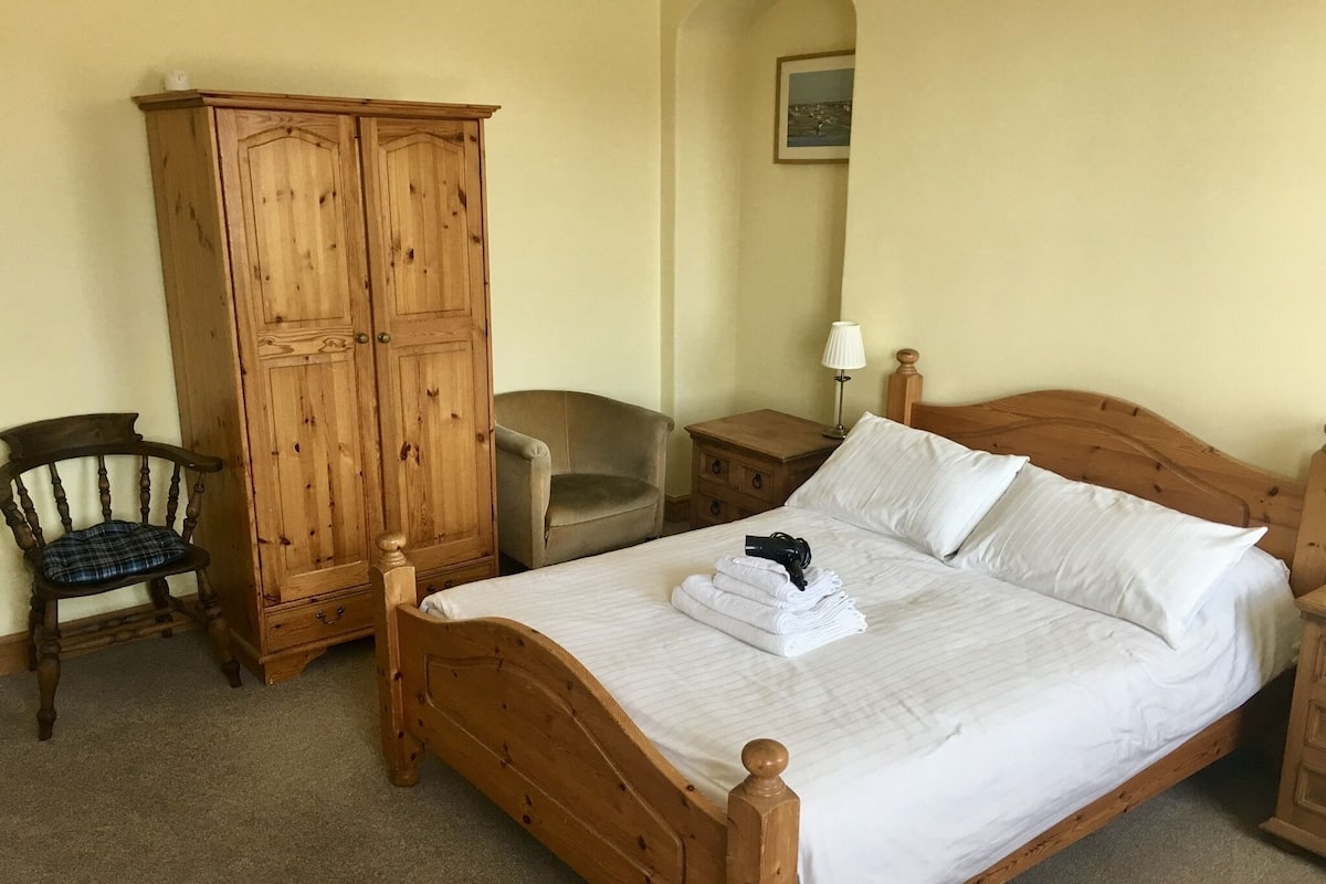 A wooden four-poster bed with white linens, a wooden wardrobe, a wooden chair, and a nightstand with a lamp.