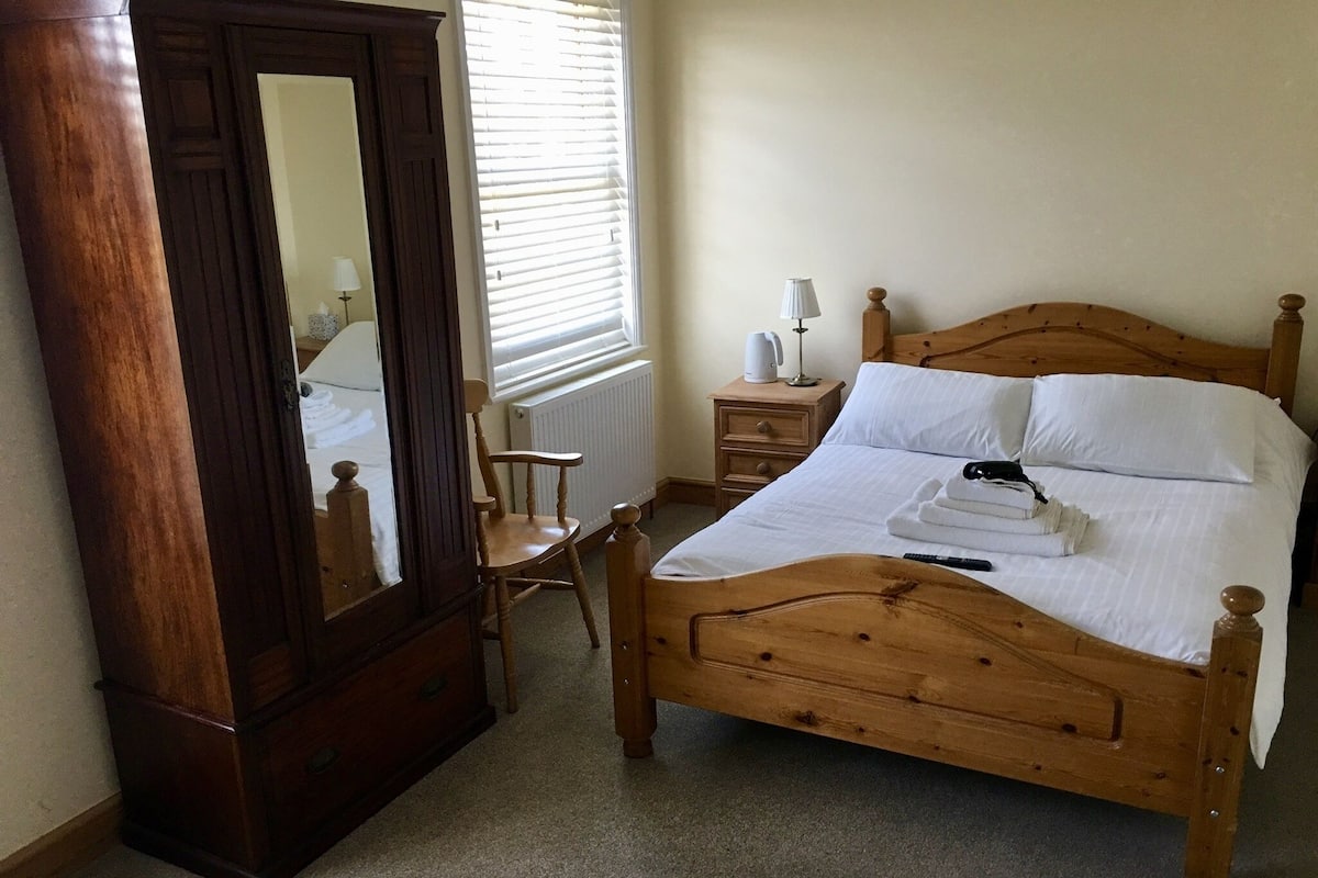 A wooden bed with white linens, a nightstand with a lamp, a wooden chair, and a large mirror in a bedroom.