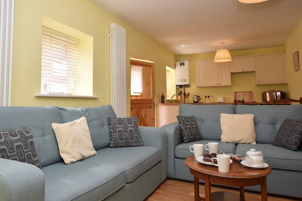 A living room with a blue sofa, a wooden coffee table with mugs and a teapot, and a kitchen area in the background.