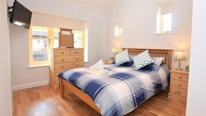 Quarto com cama, cómoda de madeira, mesas de cabeceira e televisão fixada na parede.