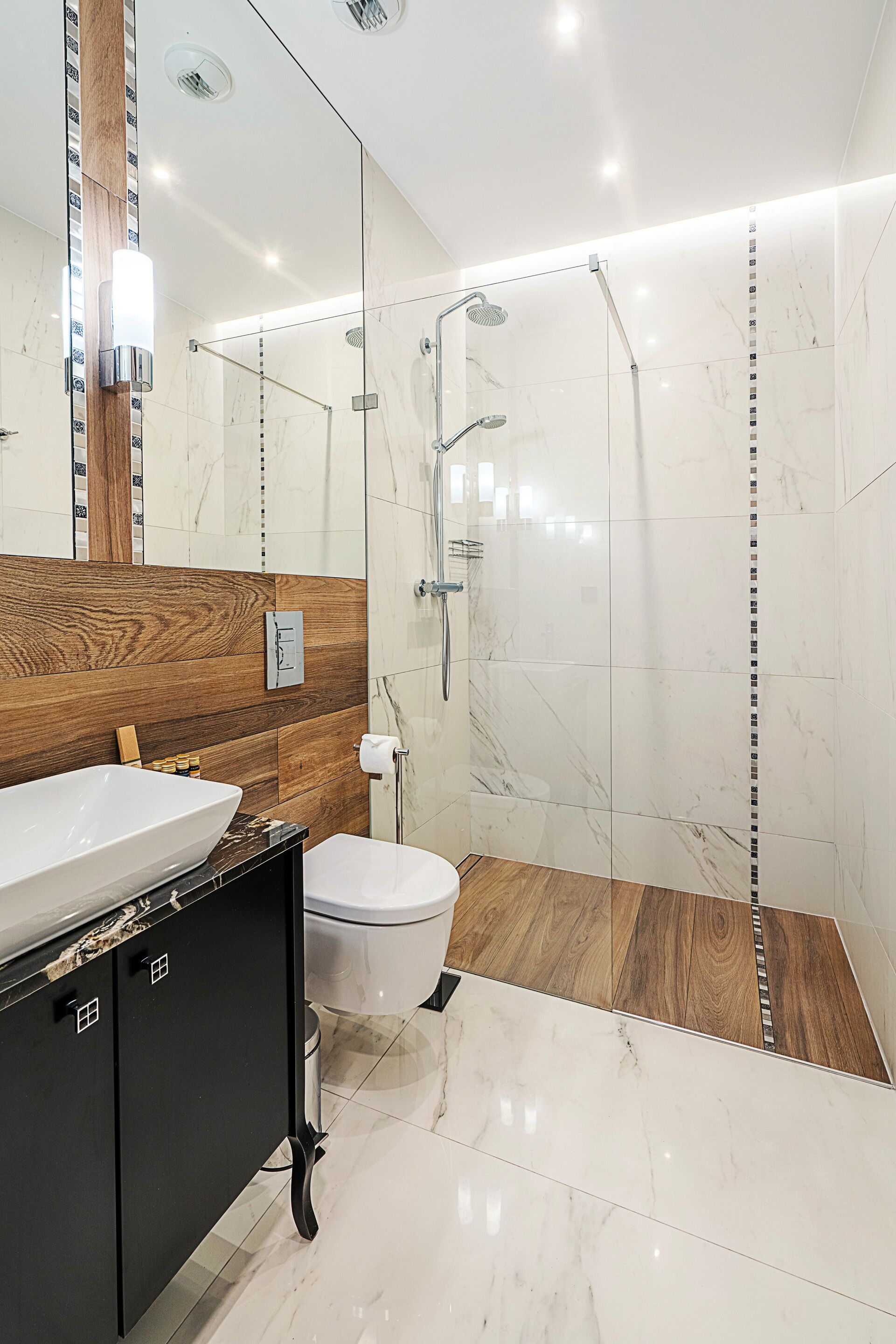 Un baño moderno con un espejo grande, un mueble de tocador de madera, un lavabo blanco, un inodoro y una zona de ducha con mampara de cristal.