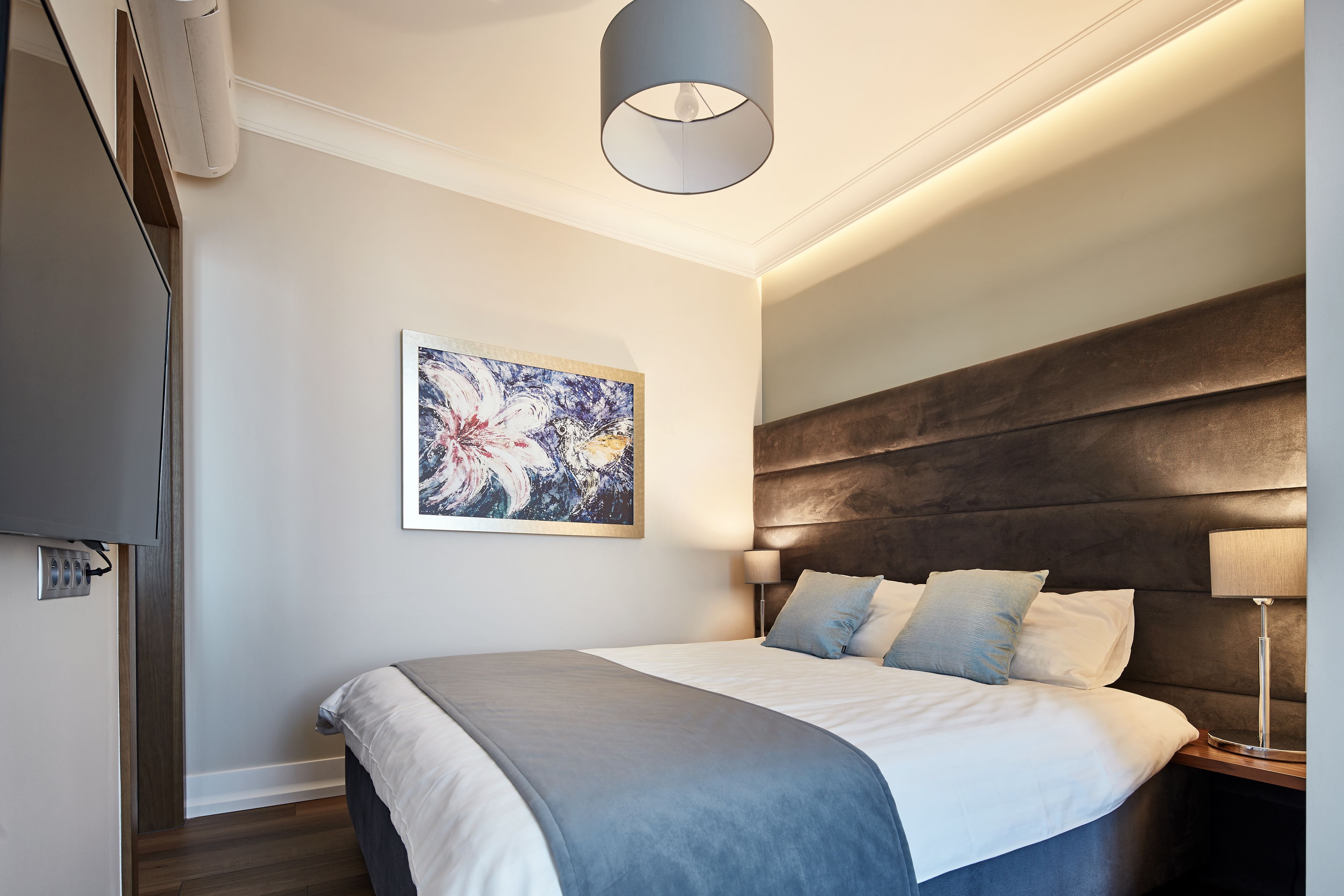 A modern hotel room with a large bed, a mounted TV, a painting on the wall, and a ceiling light fixture.