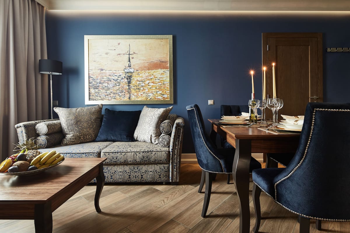 A living room with a sofa, dining table, and chairs, a painting on the wall, and a fruit bowl on the table.