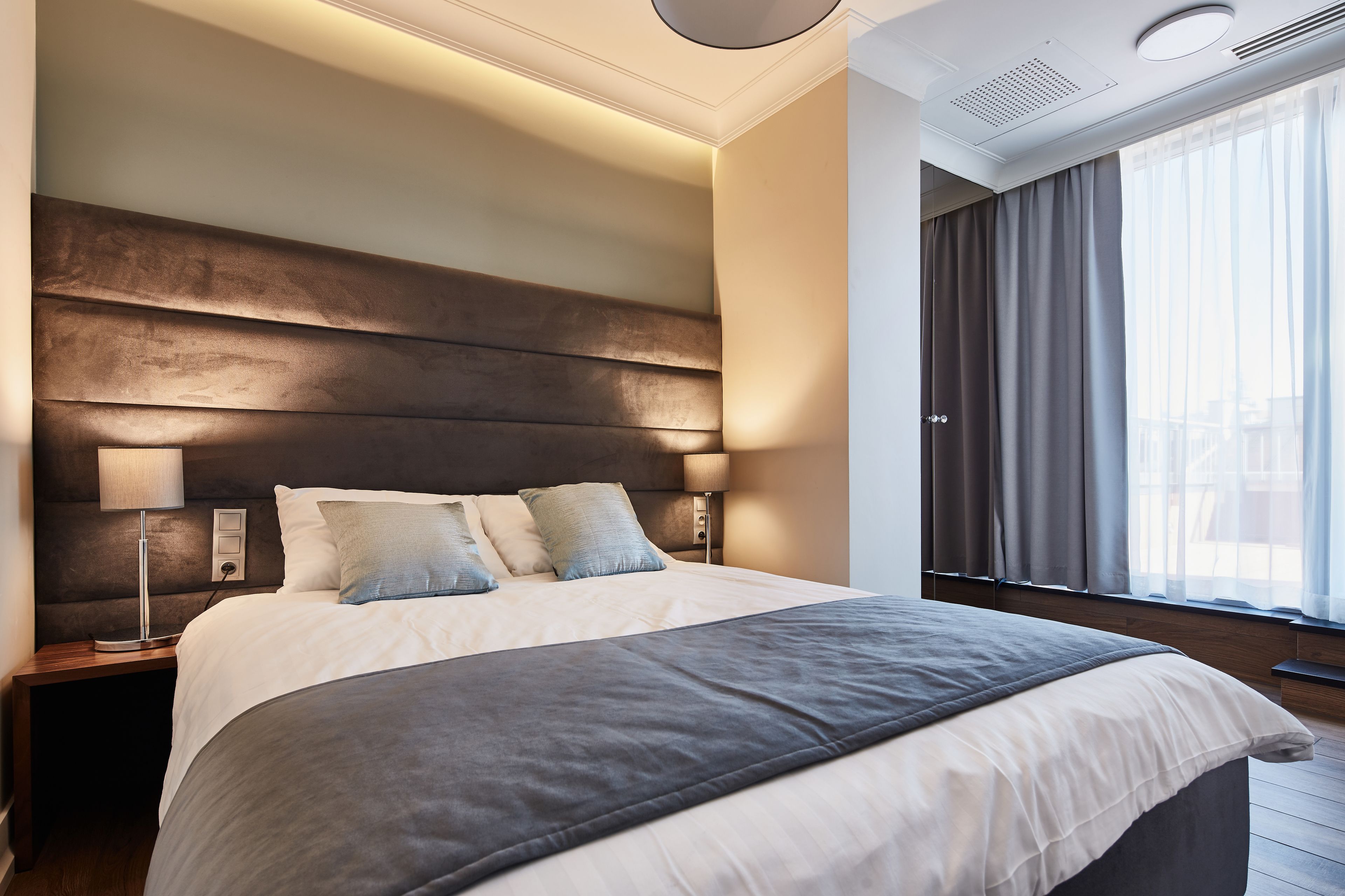 Ein modernes Hotelzimmer mit einem großen Bett, einem Nachttisch, einer Lampe und Blick ins Freie durch ein Fenster mit Vorhängen.