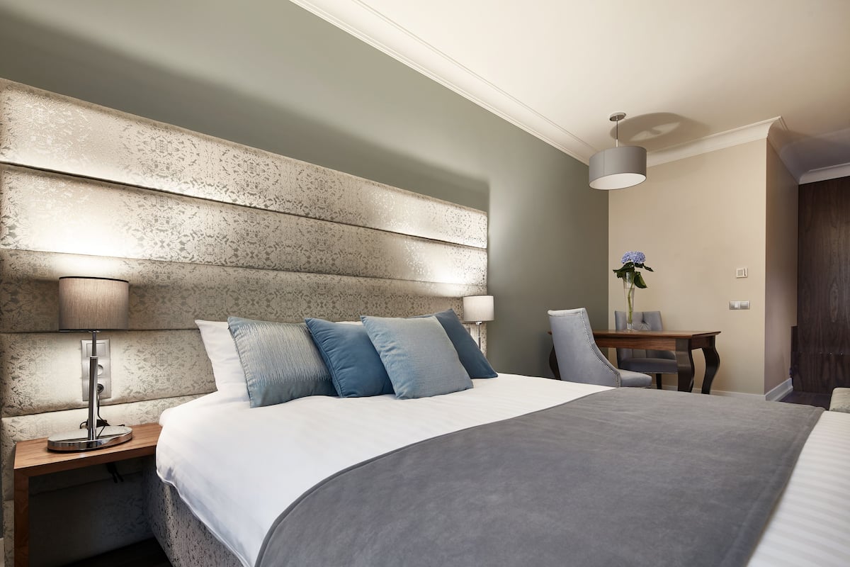 A modern hotel room with a large bed, a bedside table with a lamp, a small table with a vase of flowers, and a chair.