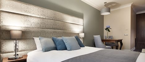 A modern hotel room with a large bed, a bedside table with a lamp, a small table with a vase of flowers, and a chair.