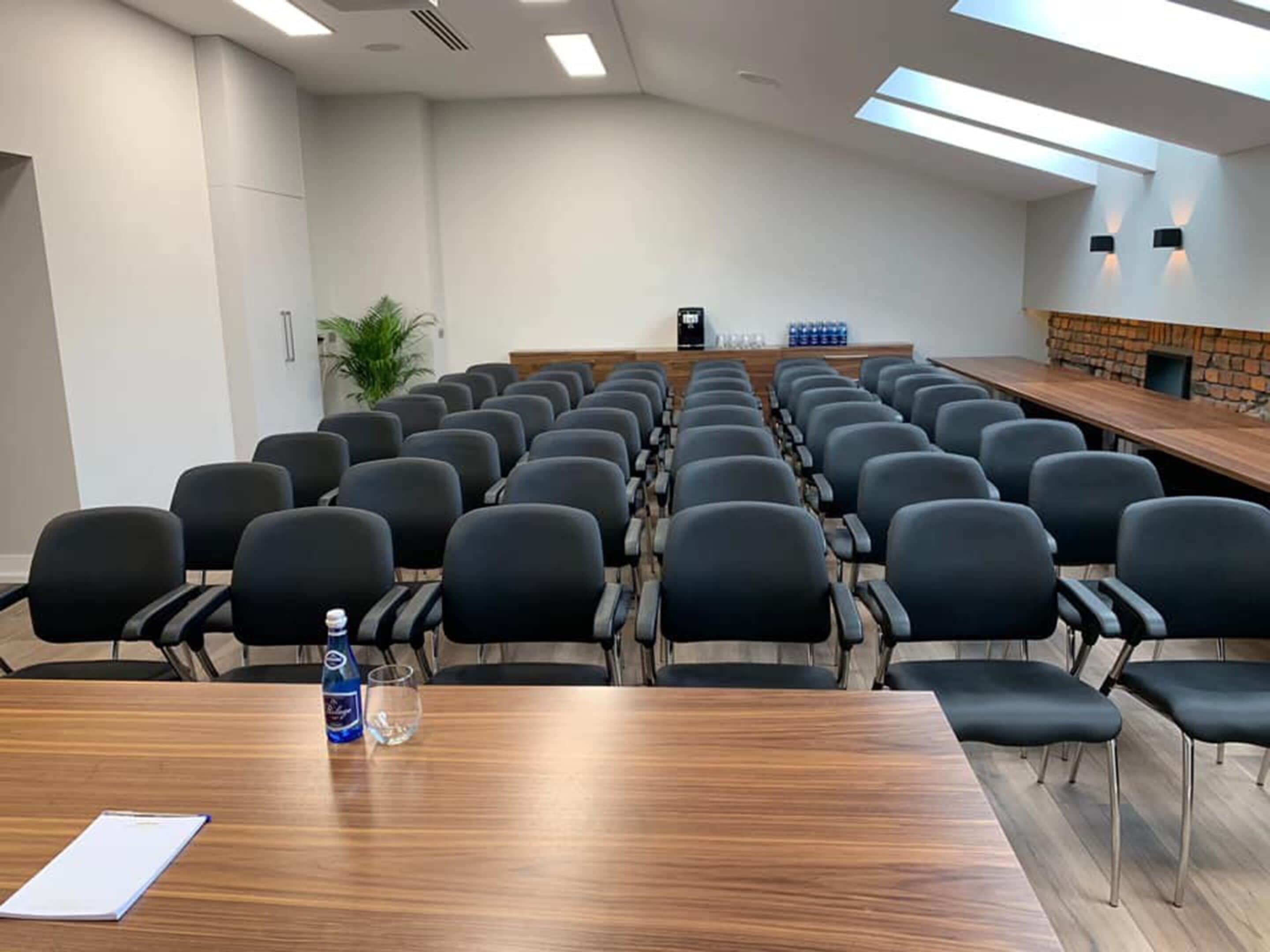 Une salle de conférence avec des rangées de chaises noires, une table en bois sur laquelle se trouvent une bouteille d’eau et un verre, et un mur décoratif en briques.