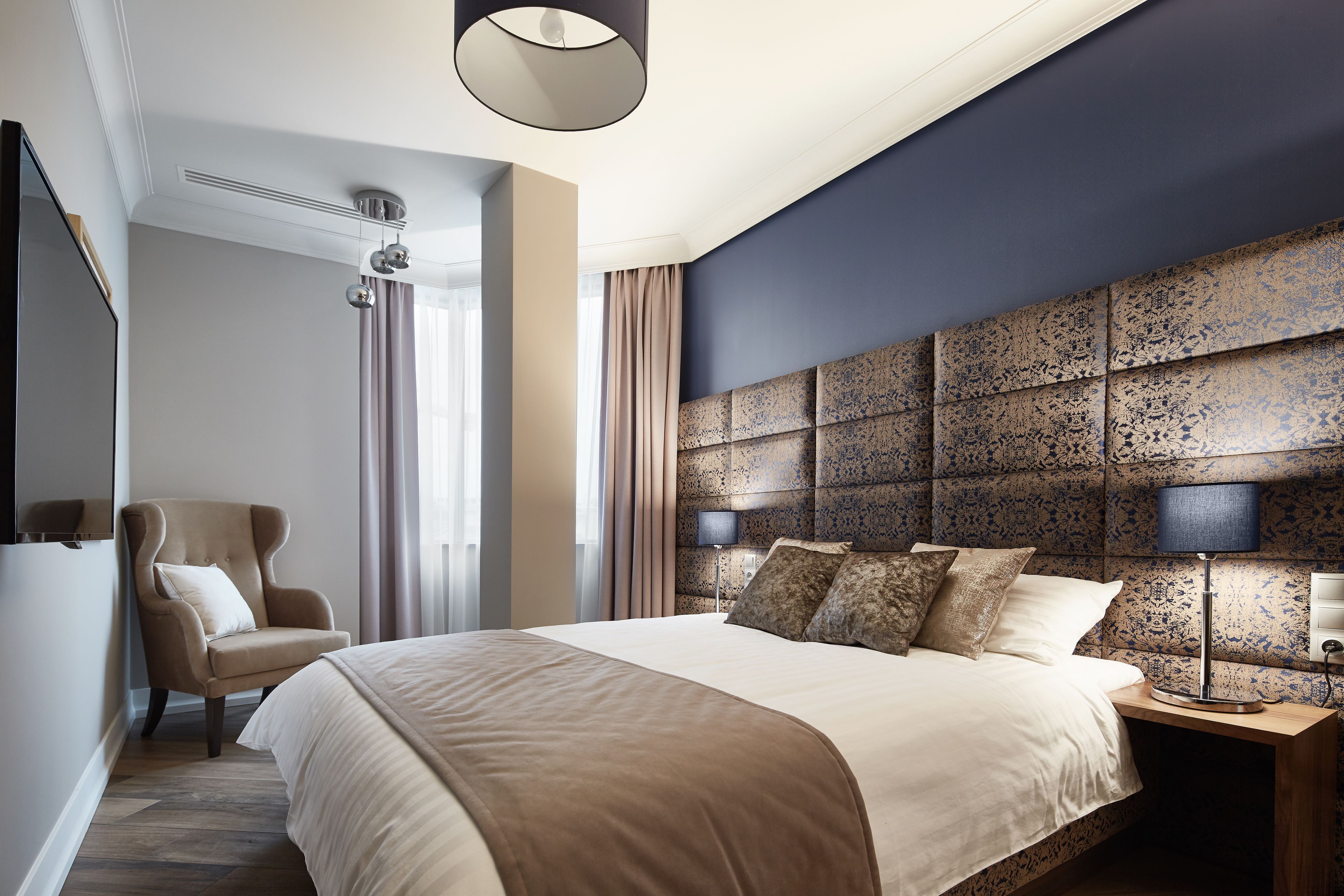 A modern hotel room with a large bed, a chair, a nightstand with a lamp, and a decorative headboard.