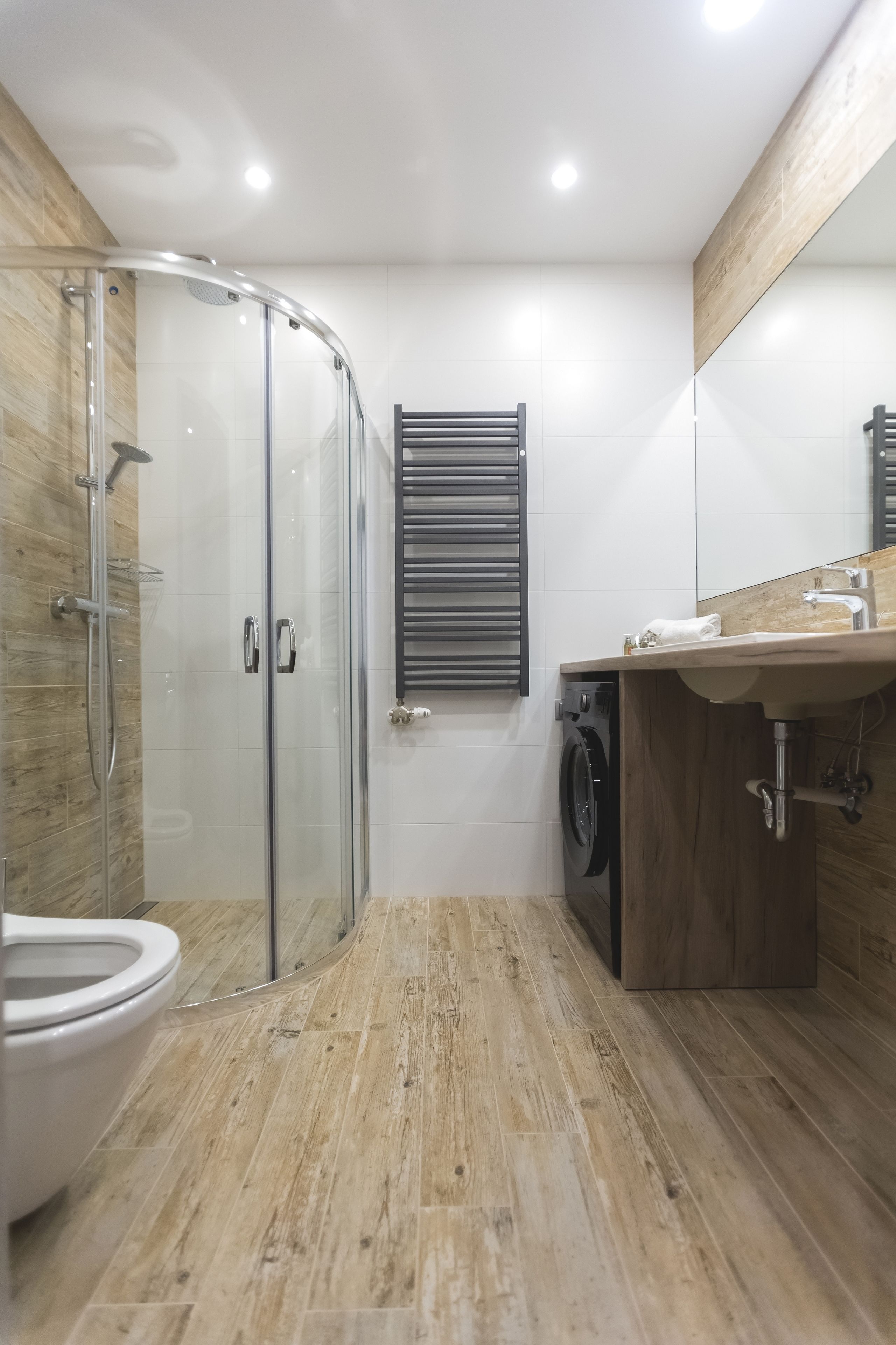 A modern bathroom with a glass shower enclosure, a wooden floor, a built-in washing machine, and a large mirror above the sink.