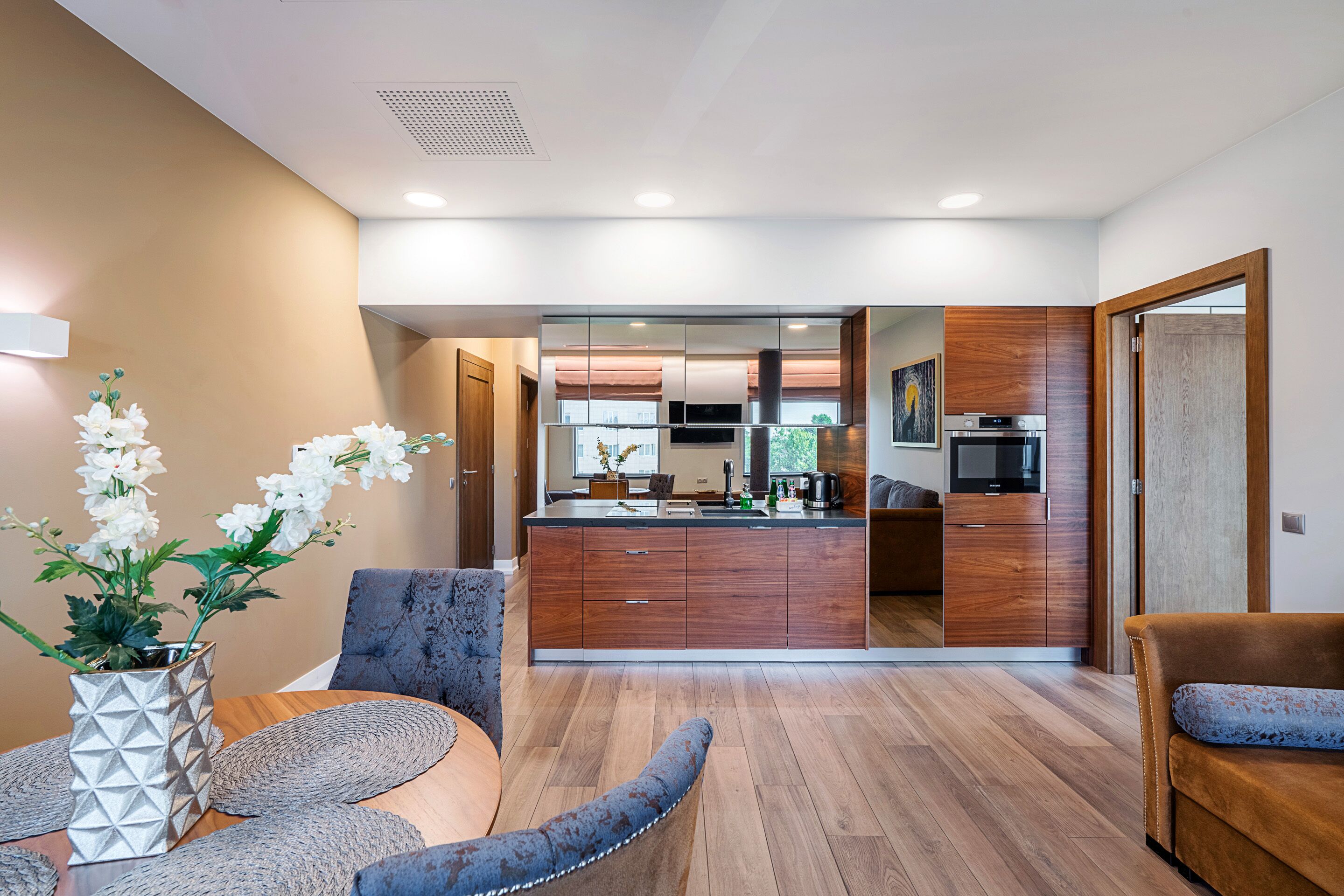 A modern kitchen with wooden cabinets, a built-in oven, and a microwave. There is a dining area with a round table and chairs, and a sofa.