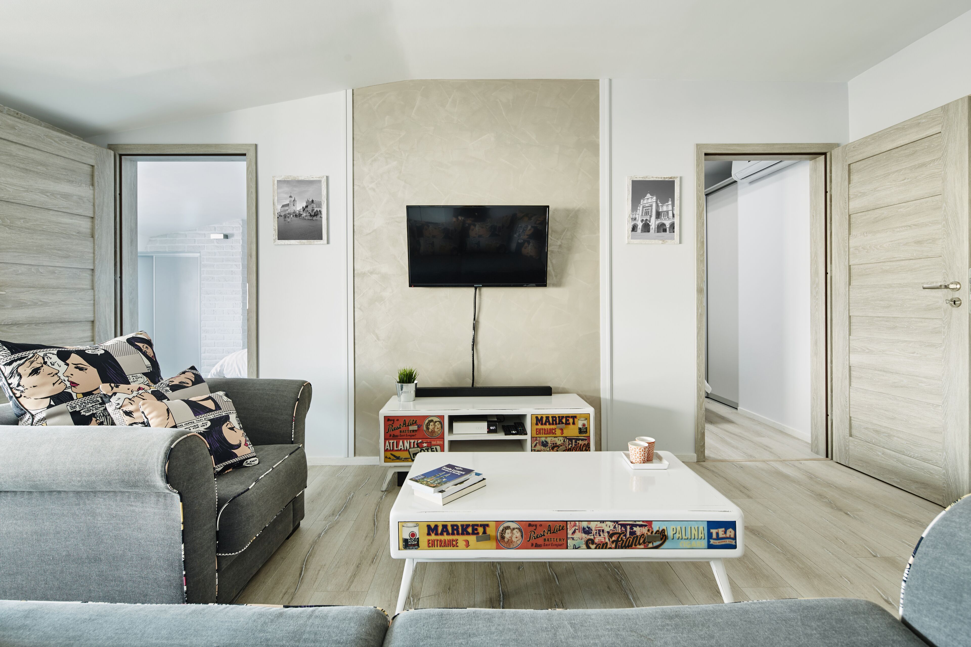 A modern living room with a grey sofa, a white coffee table with magazine covers, a flat-screen TV on the wall, and a small plant on a white console table.