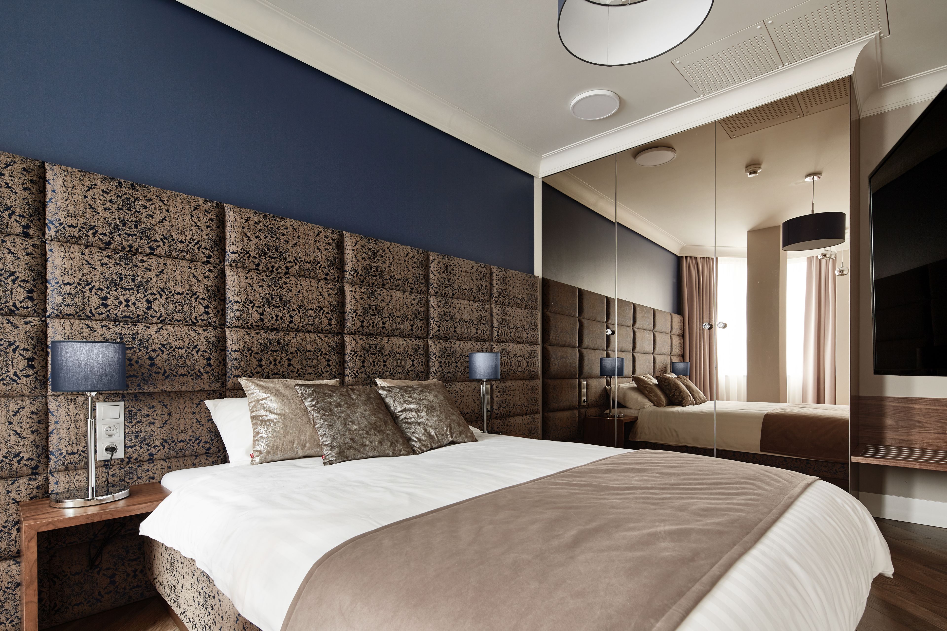 A modern bedroom with a large bed, a headboard with a textured pattern, bedside lamps, and a mirror.