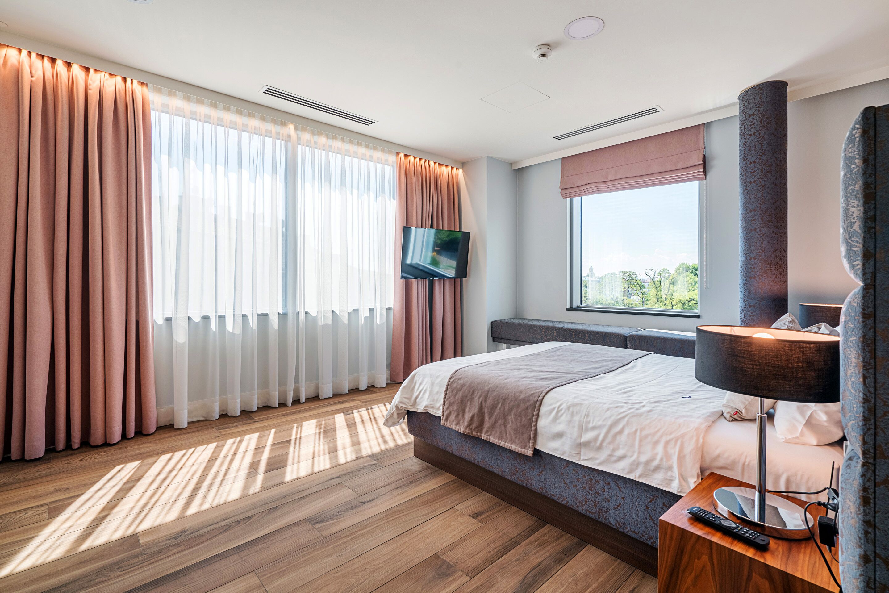 Een moderne hotelkamer met een groot bed, een flatscreen televisie, een nachtkastje met lamp en een raam met gordijnen.