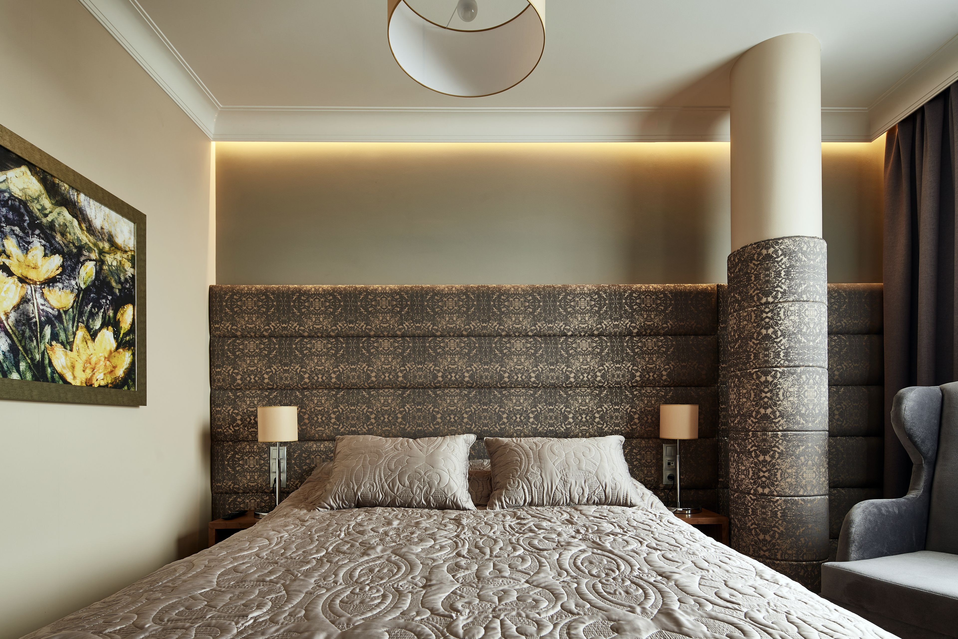 A hotel room with a large bed, two bedside lamps, a headboard with a textured pattern, a painting on the wall, and a large pillar.
