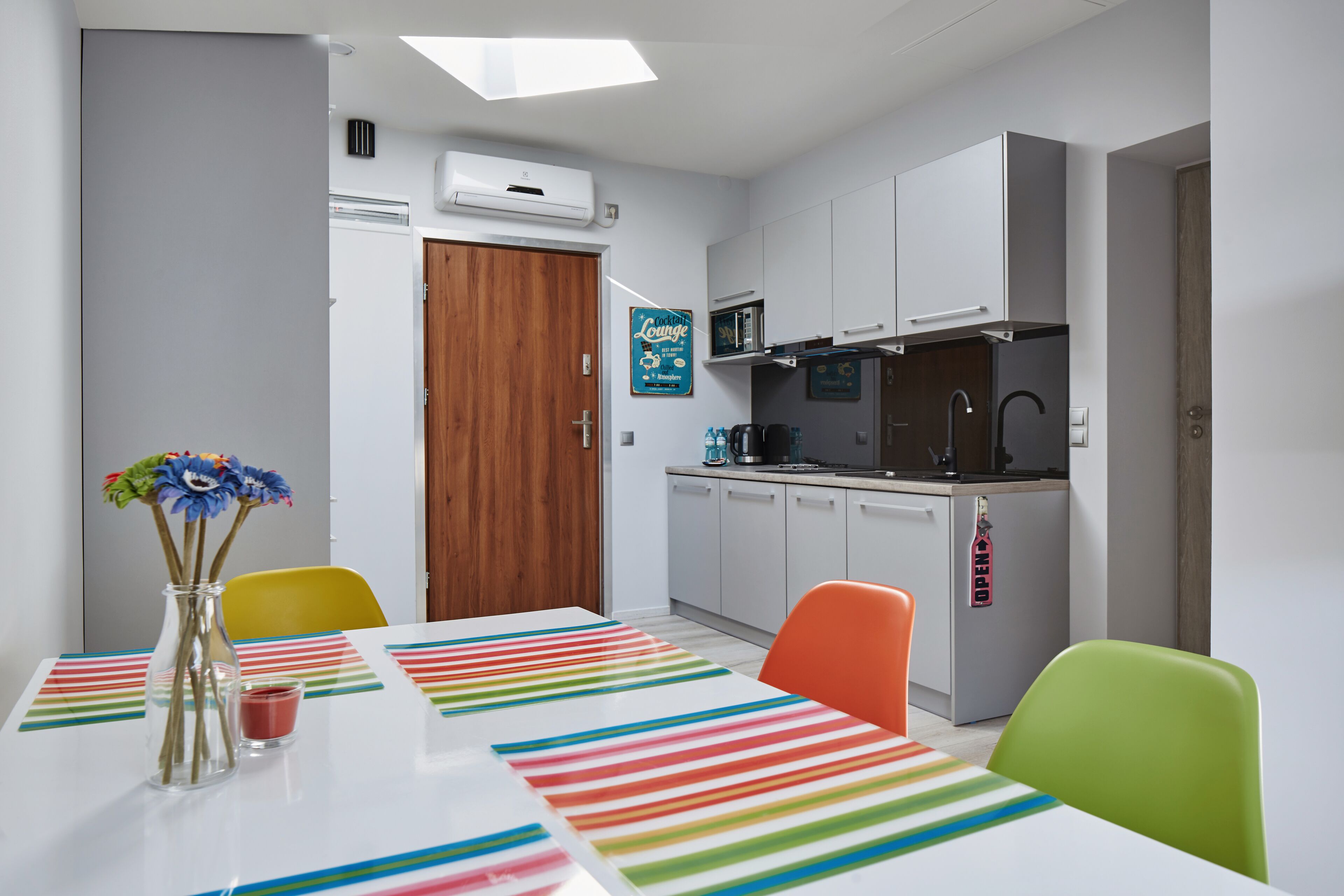 A modern kitchen with white cabinets, a microwave, and a coffee machine. There is a dining table with colorful striped placemats and a vase with flowers.