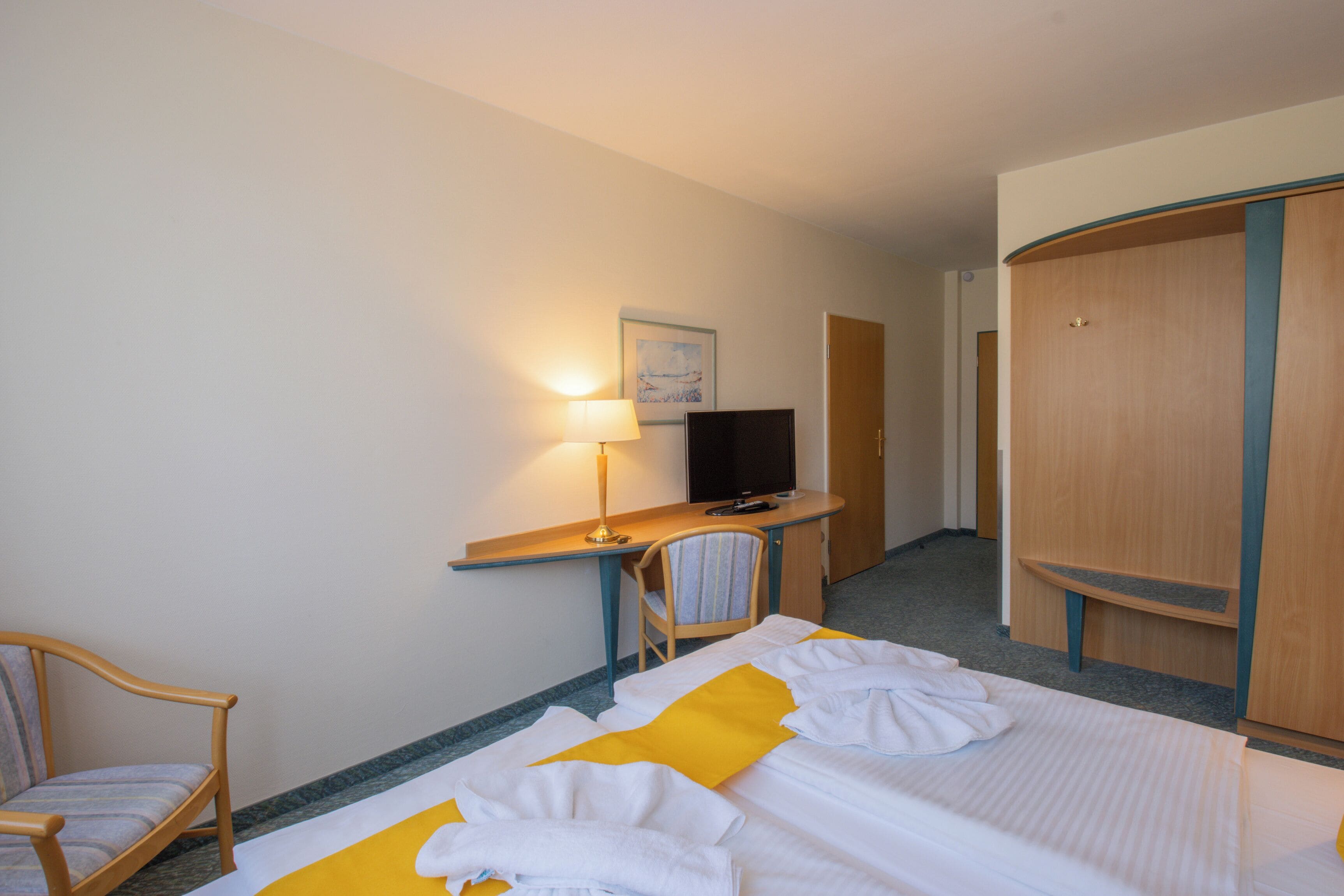 Una habitación de hotel con cama, escritorio con lámpara, silla, televisor y armario.