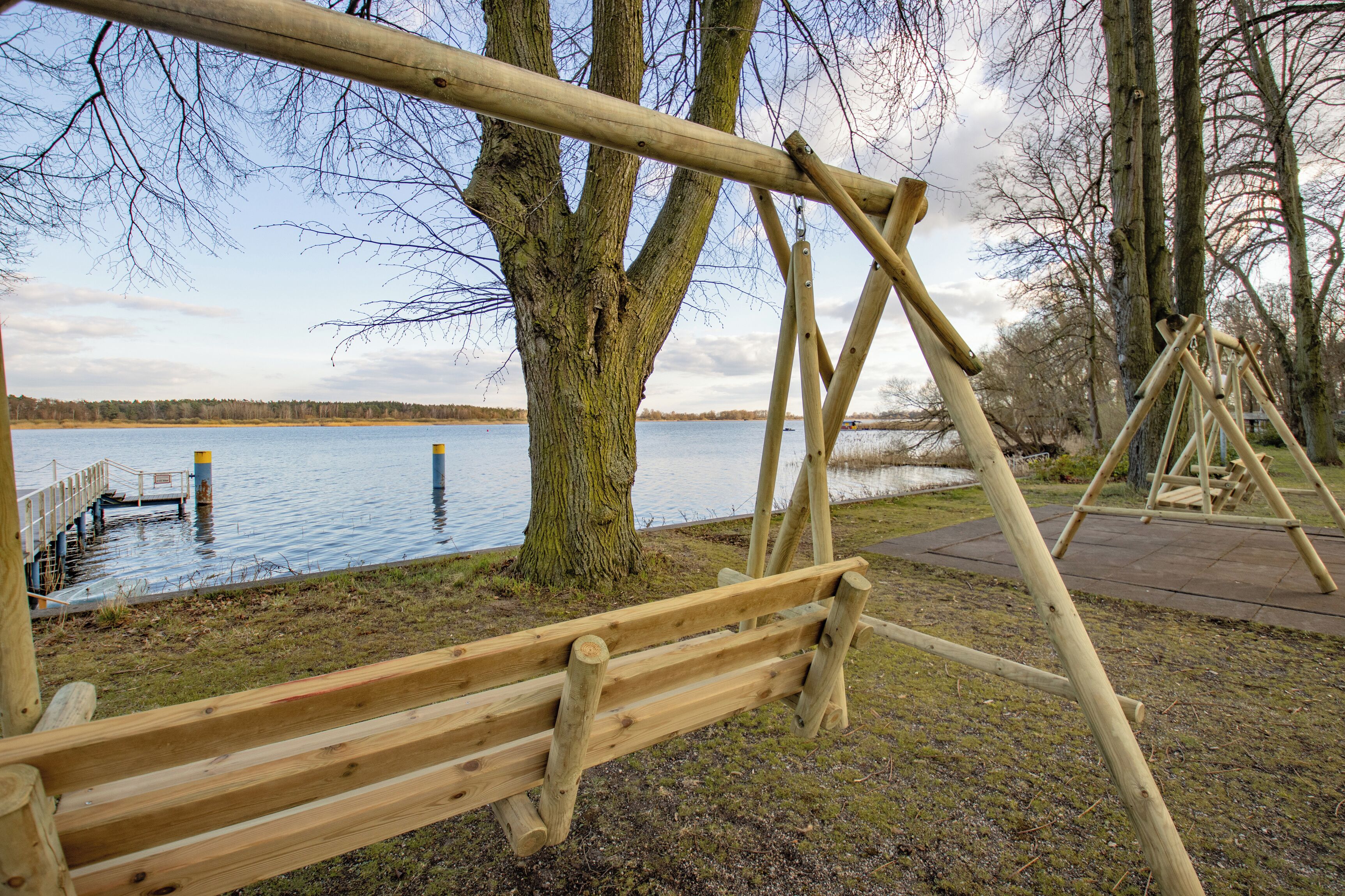 Een houten schommelset bij een meer, met een steiger en een boom op de voorgrond.