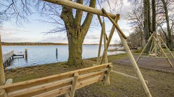 Een houten schommelset bij een meer, met een steiger en een boom op de voorgrond.