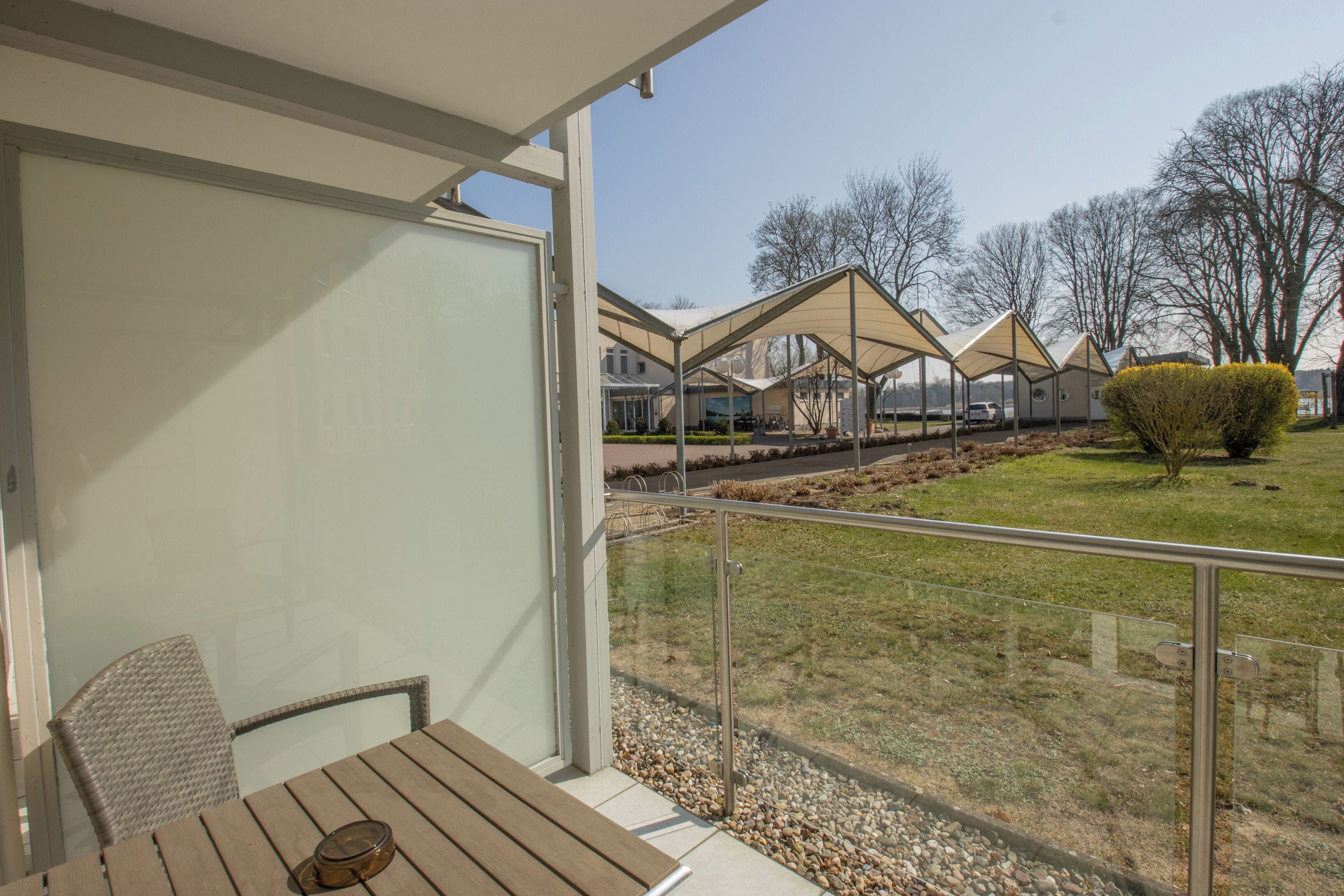 Een balkon met een houten tafel en stoelen, een glazen hekwerk en uitzicht op een woonwijk met huizen en gazons.