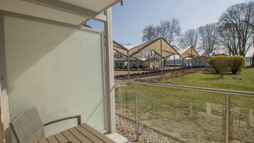 Een balkon met een houten tafel en stoelen, een glazen hekwerk en uitzicht op een woonwijk met huizen en gazons.