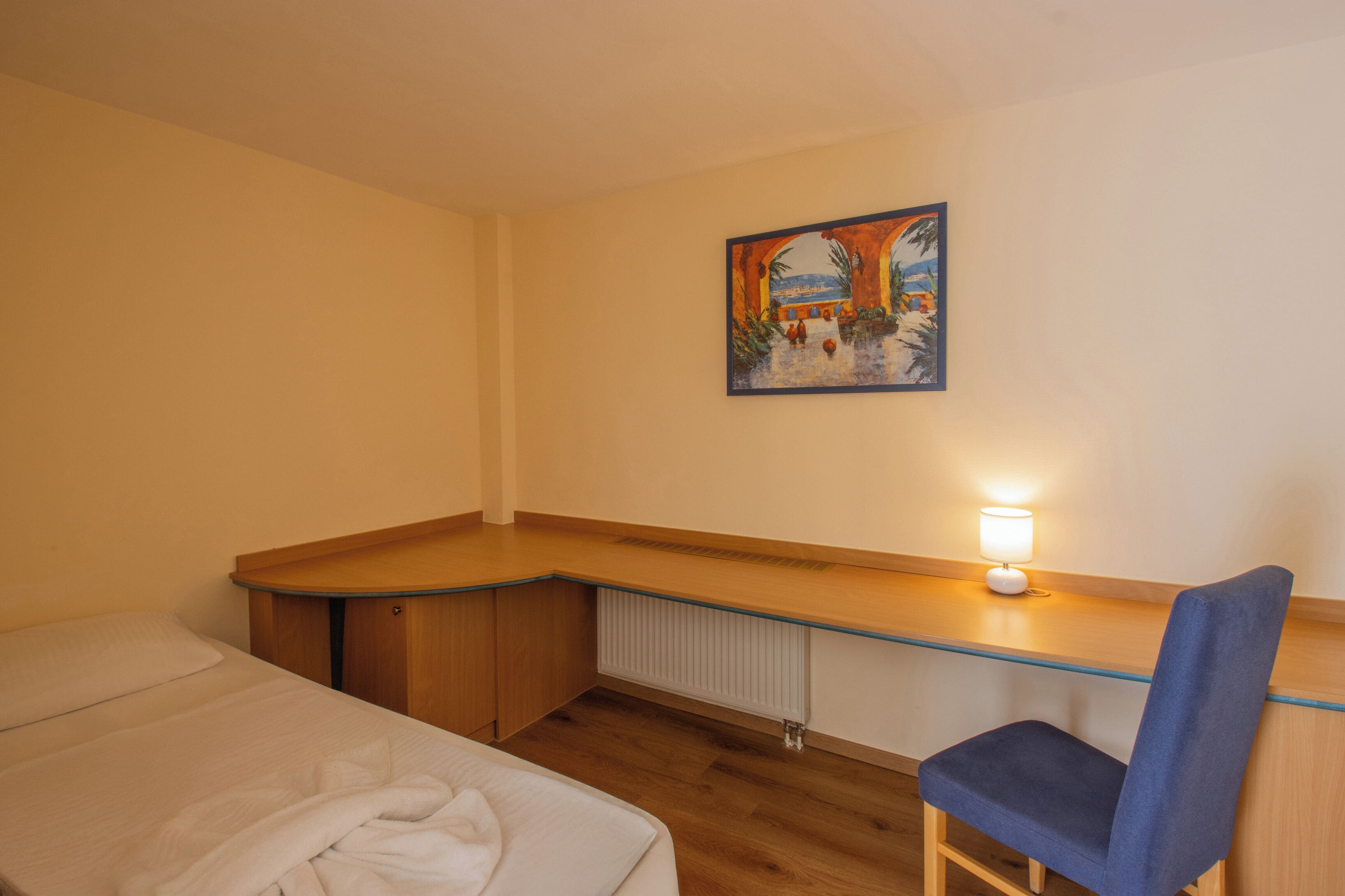 Een slaapkamer met een bed, een bureau met een stoel, een radiator en een schilderij aan de muur.