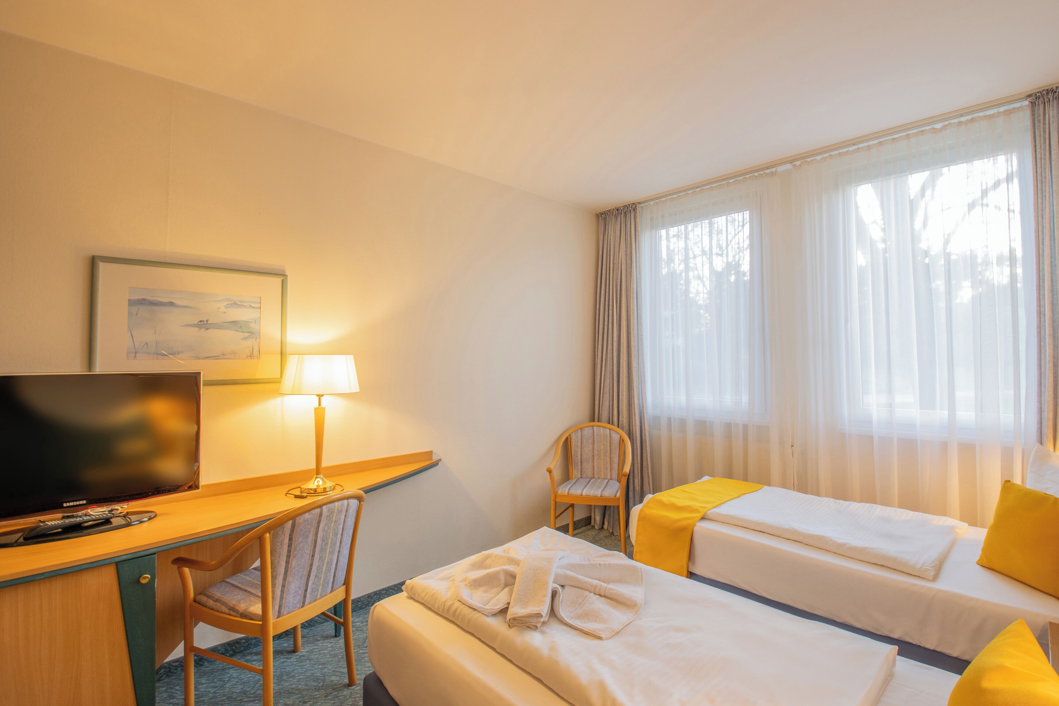 Een hotelkamer met een bureau, televisie, twee bedden, een stoel en een raam met gordijnen.