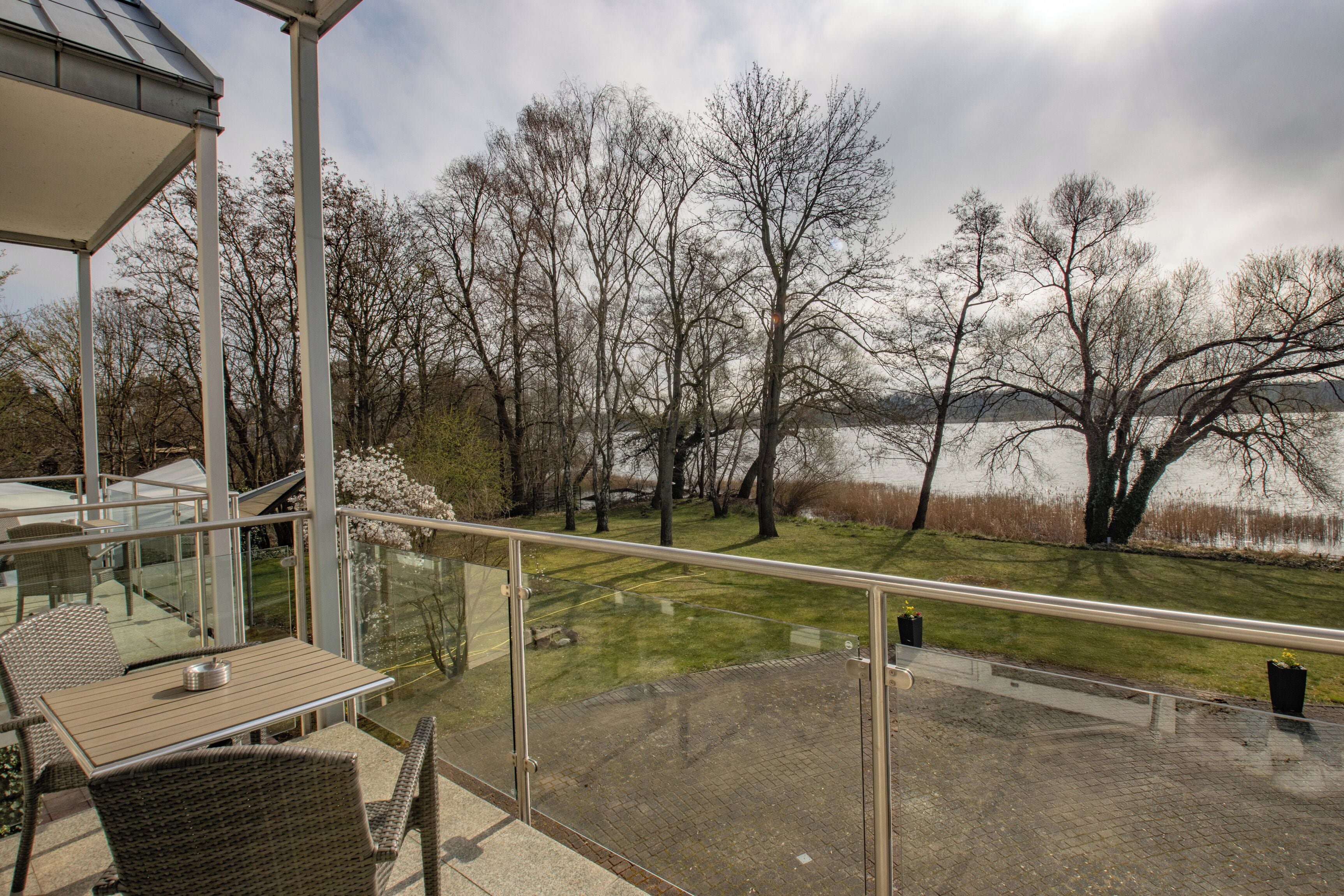 A balcony with wicker furniture, a glass railing, and a view of a lake and trees.