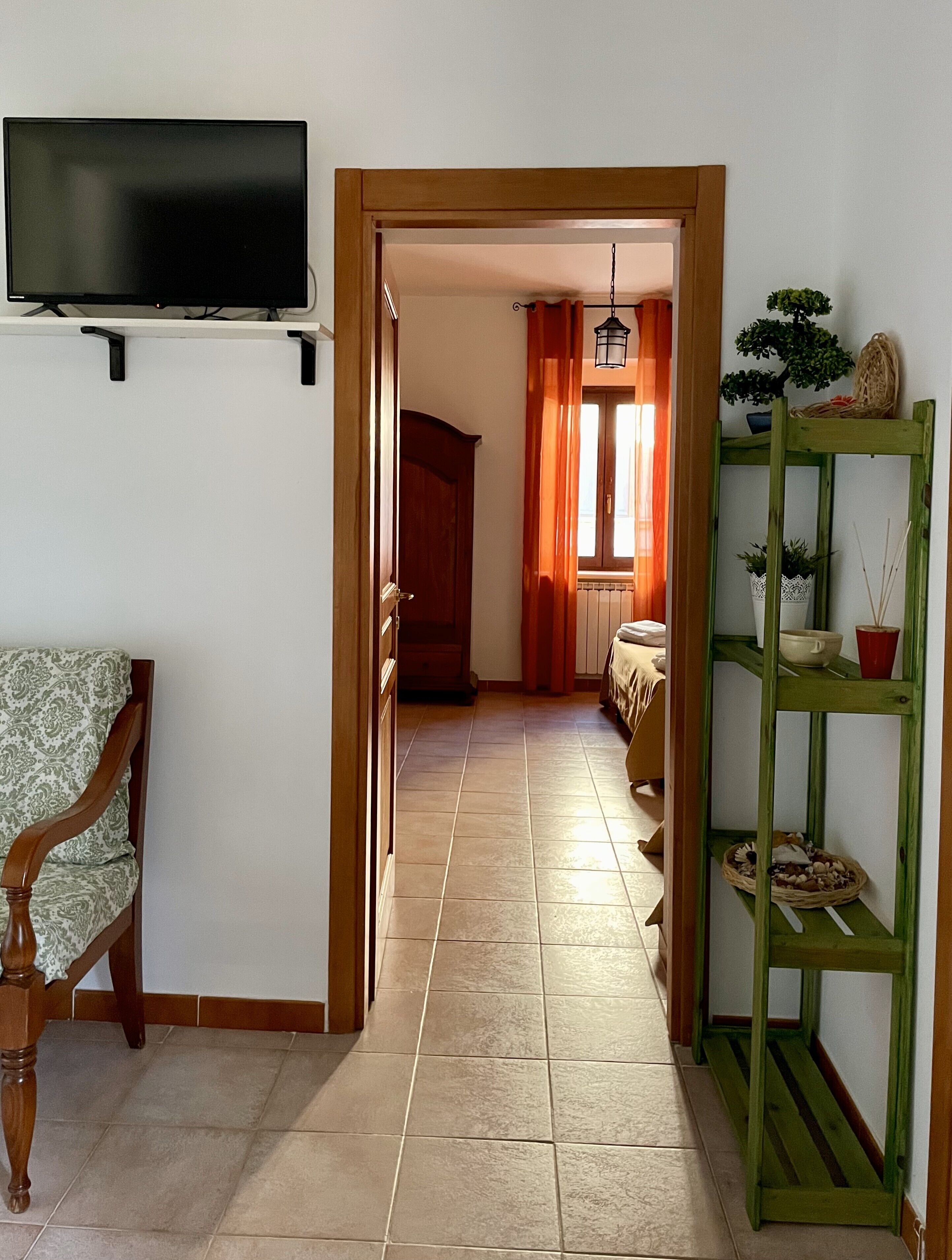 Un pasillo con un televisor montado en la pared, una silla de madera, un estante verde con plantas y un suelo de baldosas.