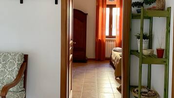 Un pasillo con un televisor montado en la pared, una silla de madera, un estante verde con plantas y un suelo de baldosas.