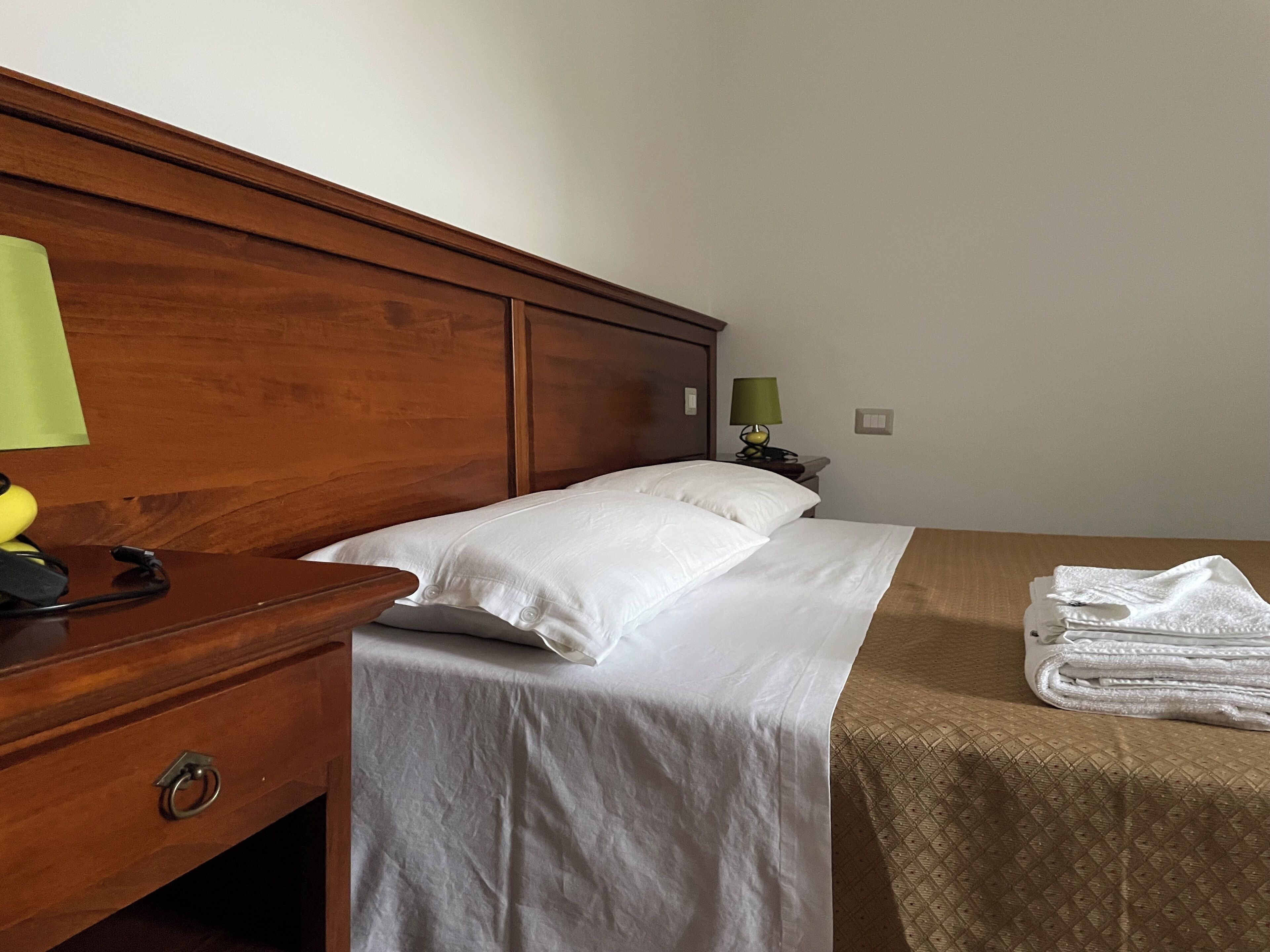 Una habitación de hotel con cabecera de madera, una cama con ropa blanca, una mesita de noche con lámpara y una toalla doblada sobre la cama.