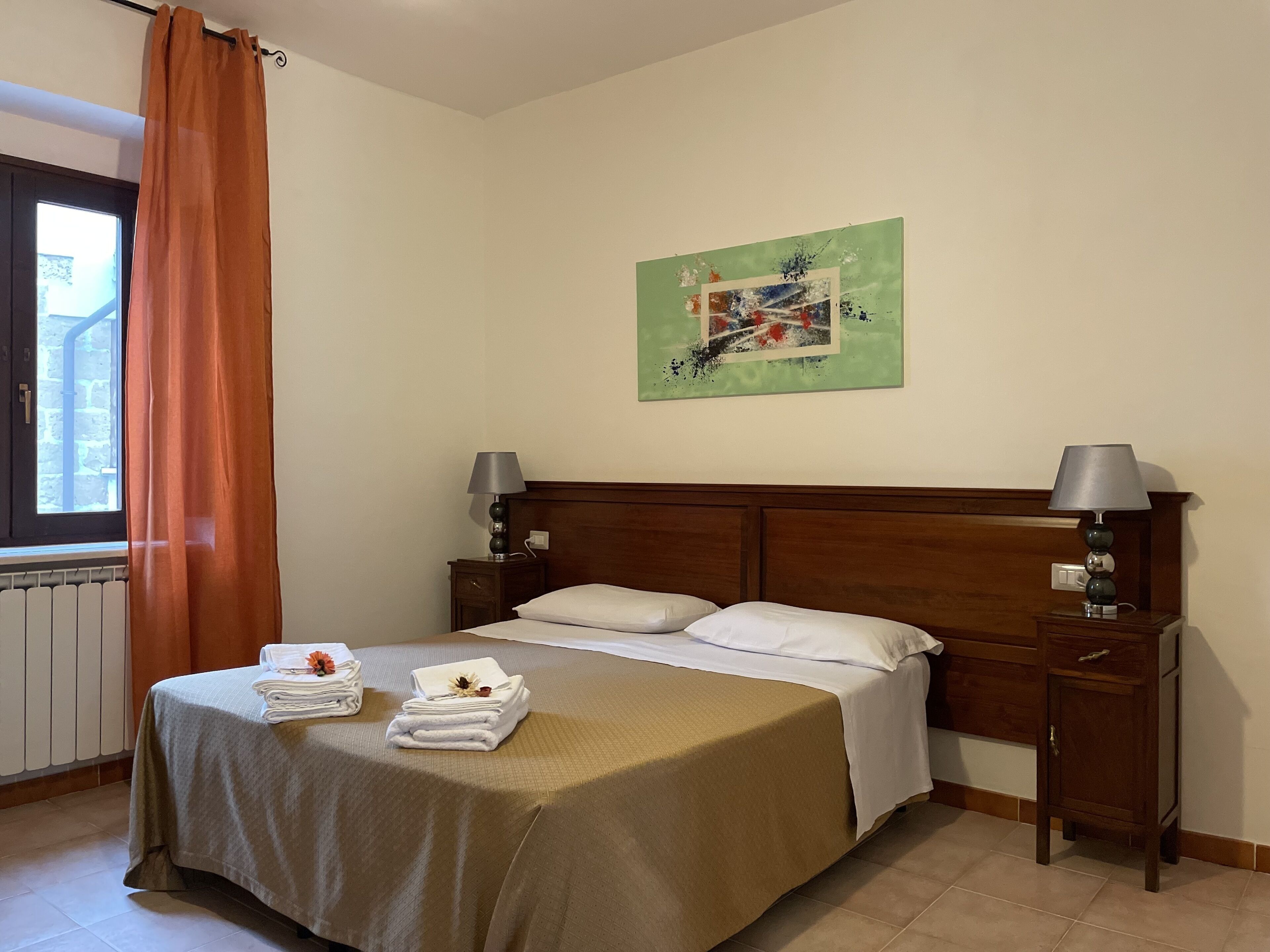 A neatly made bed with a wooden headboard, two bedside tables with lamps, a window with orange curtains, and a painting on the wall.