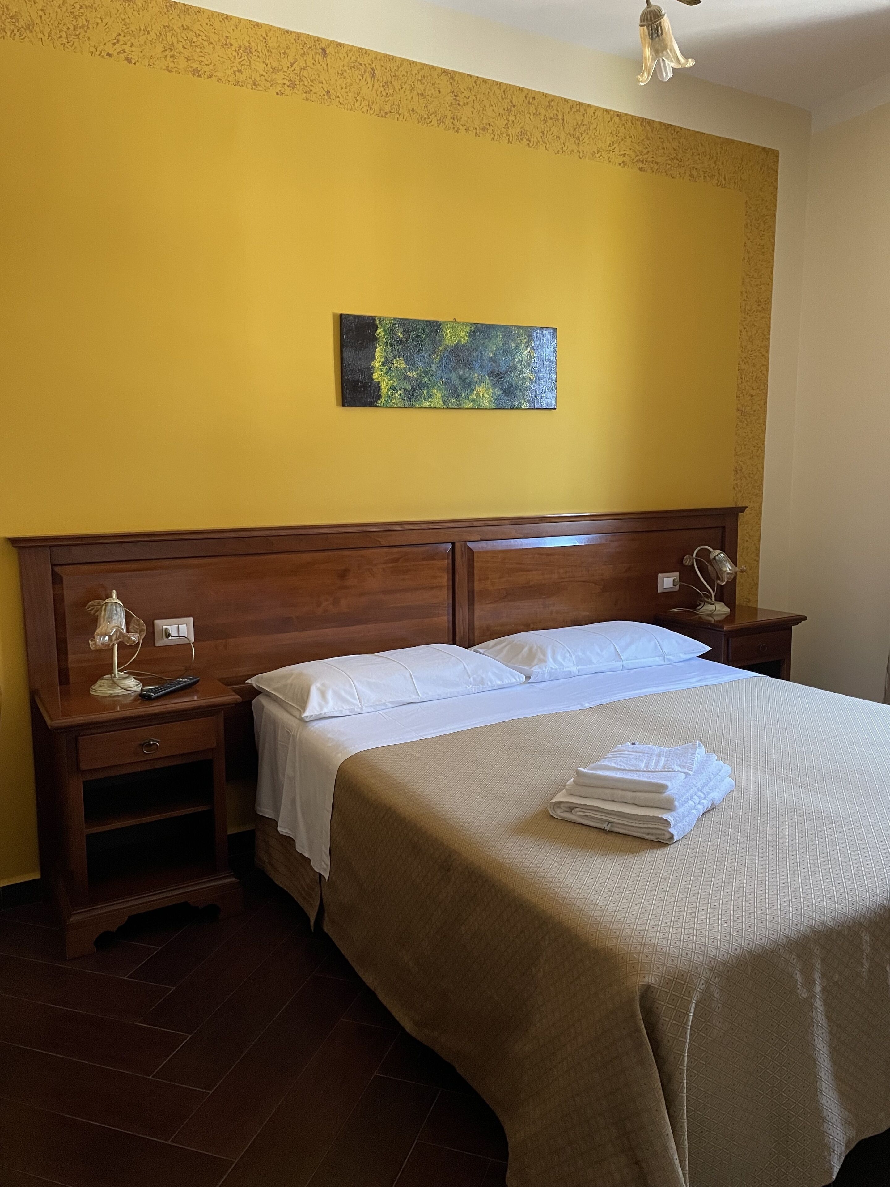 Una camera d'albergo con una testiera in legno, un letto con lenzuola bianche, un comodino con una lampada e un quadro appeso al muro.