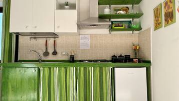Una cocina con armarios blancos, una encimera verde y un refrigerador blanco. Hay dos repisas sobre la encimera, una con una planta y la otra con utensilios de cocina.