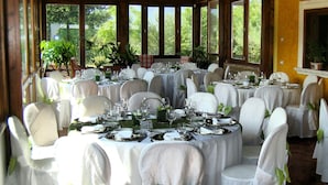 Un restaurante con mesas redondas dispuestas para comer, sillas blancas decoradas con lazos y grandes ventanales que dan a zonas verdes.