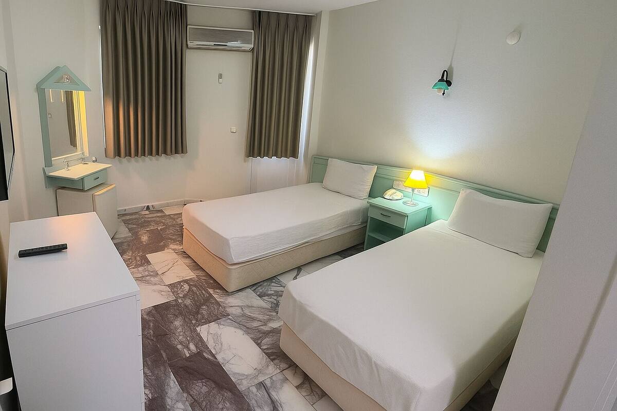A hotel room with two single beds, a small bedside table with a lamp, a mirror, and an air conditioning unit.