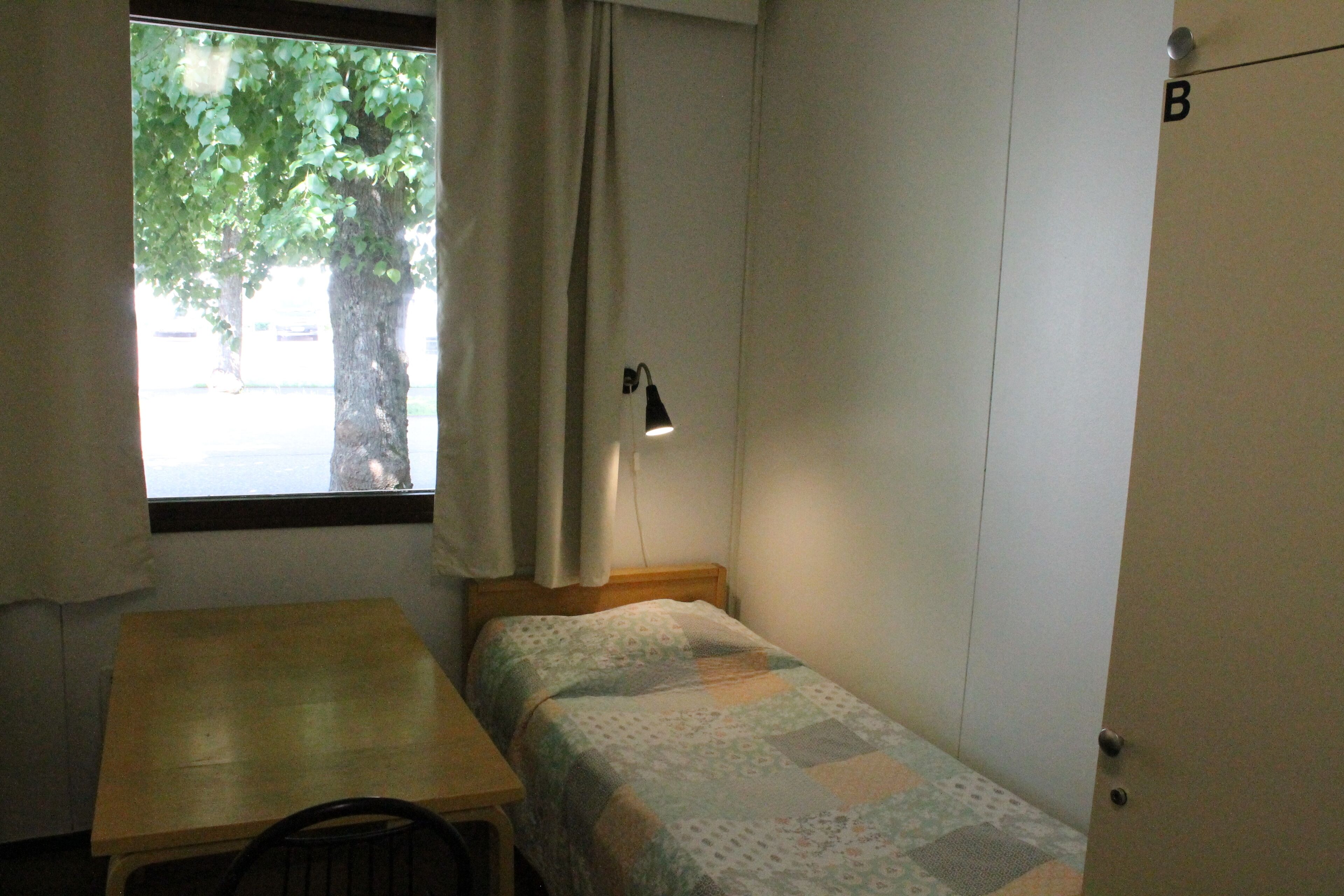 A small, single-bed room with a wooden table, a window with curtains, a desk lamp, and a door with a letter 'B' on it.