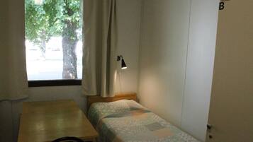 A small, single-bed room with a wooden table, a window with curtains, a desk lamp, and a door with a letter 'B' on it.
