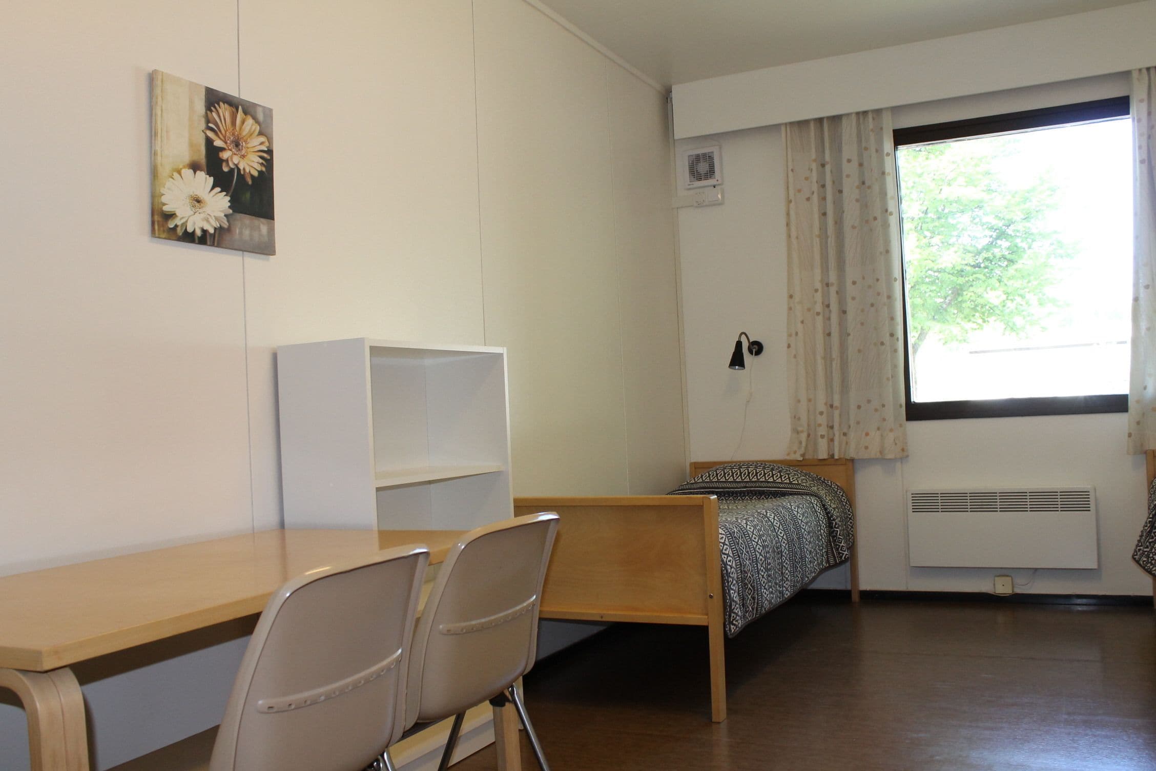 Un dormitorio con cama de madera, un escritorio, dos sillas, una ventana con cortinas y un radiador.