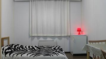 Una habitación pequeña con cama individual, colcha de estampado de cebra, una mesita con mantel, una lámpara roja y un radiador blanco.
